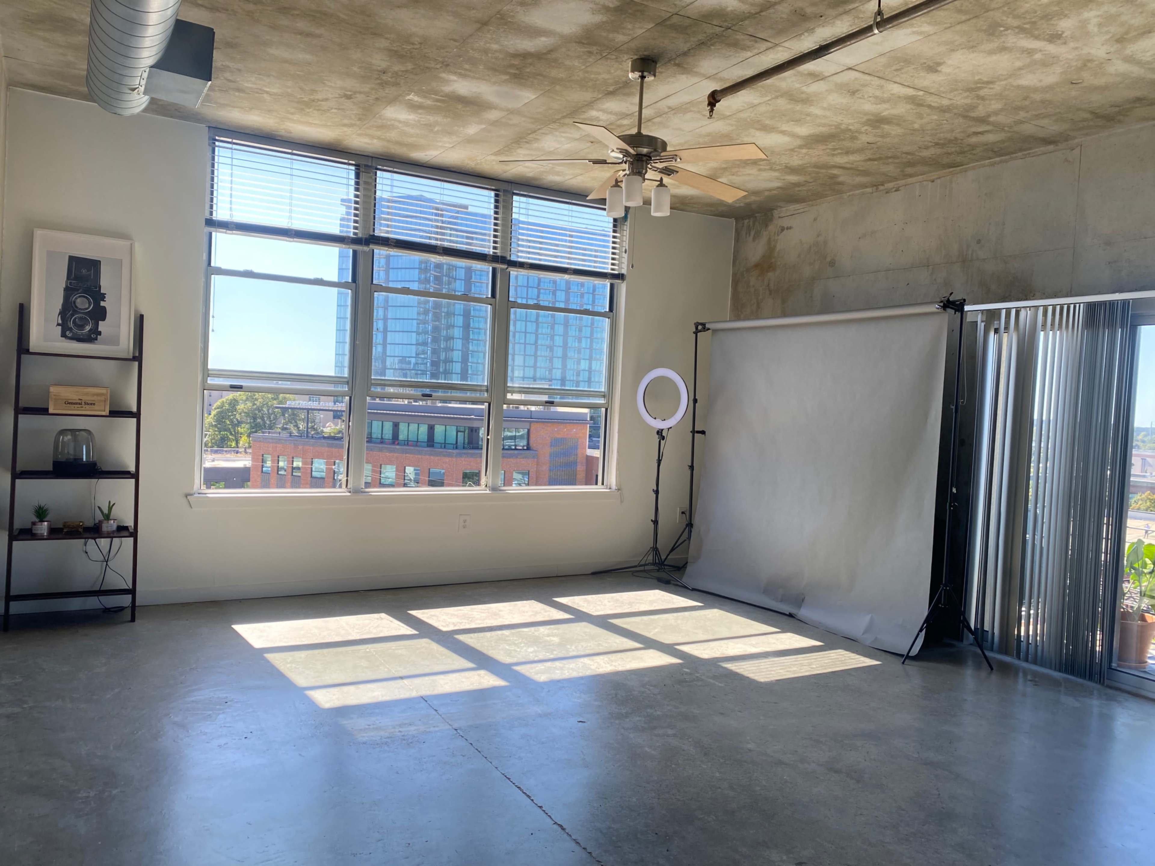 A spacious, well-lit room features large windows, a ceiling fan, and a backdrop setup against a concrete wall.