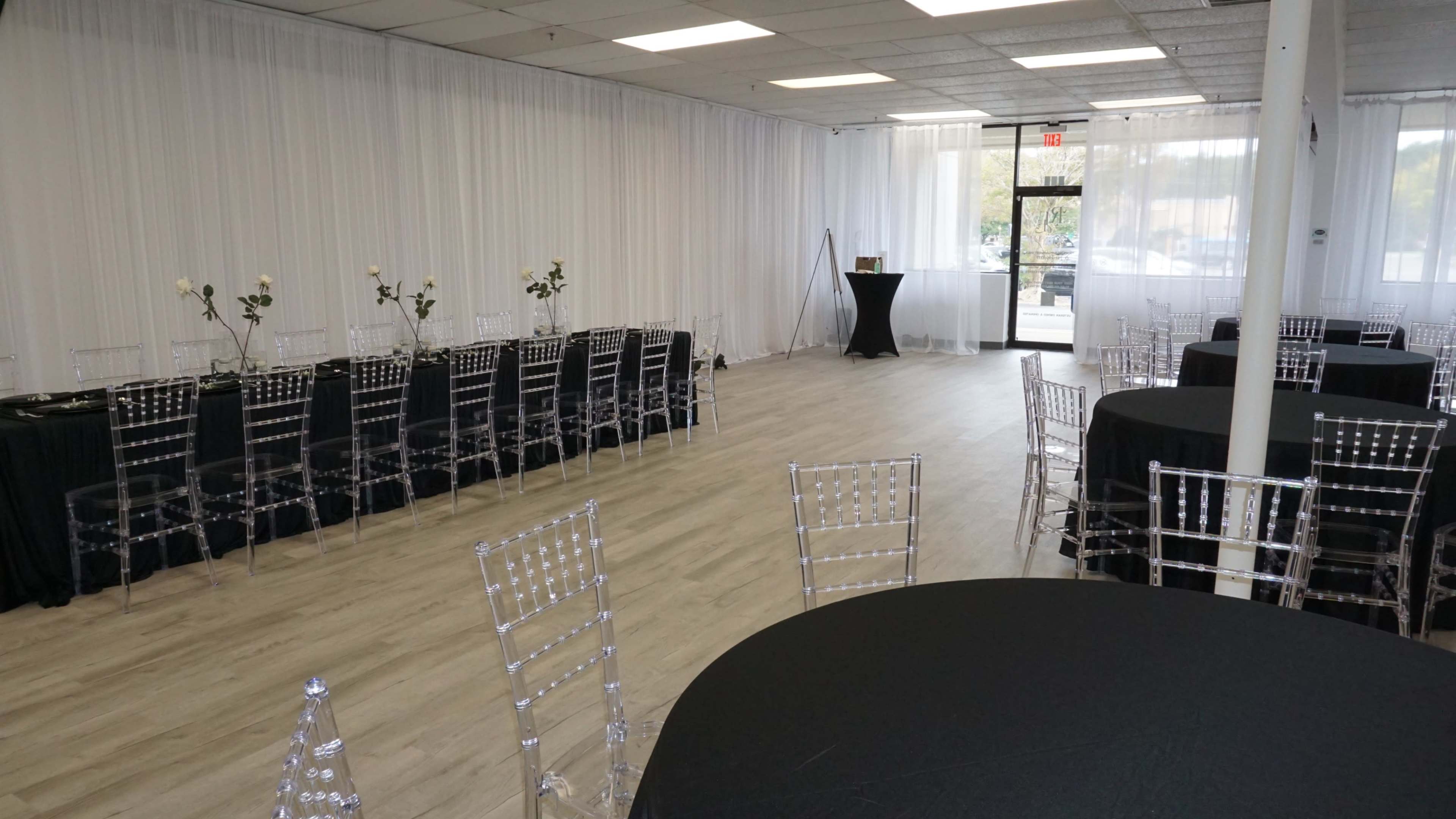 The image shows a spacious event venue with tables covered in black cloth and clear chairs, illuminated by natural light filtering through large windows.