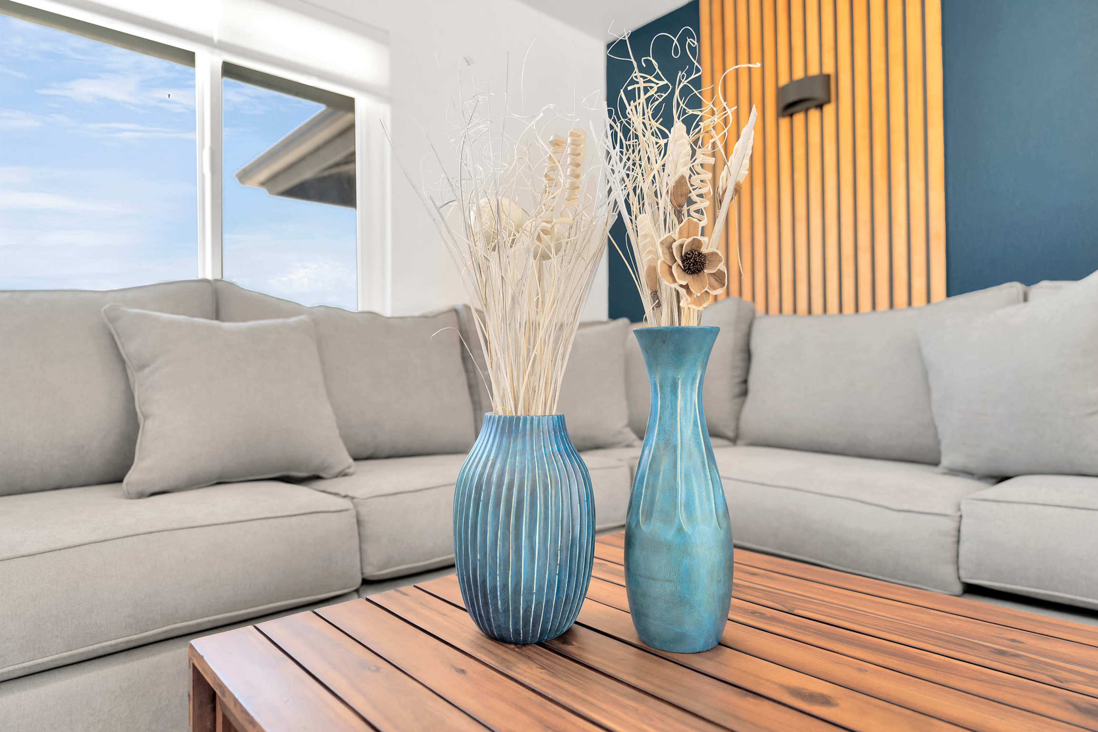 The image shows two decorative vases with dried flowers on a wooden coffee table in a light-filled living room with a gray sofa.