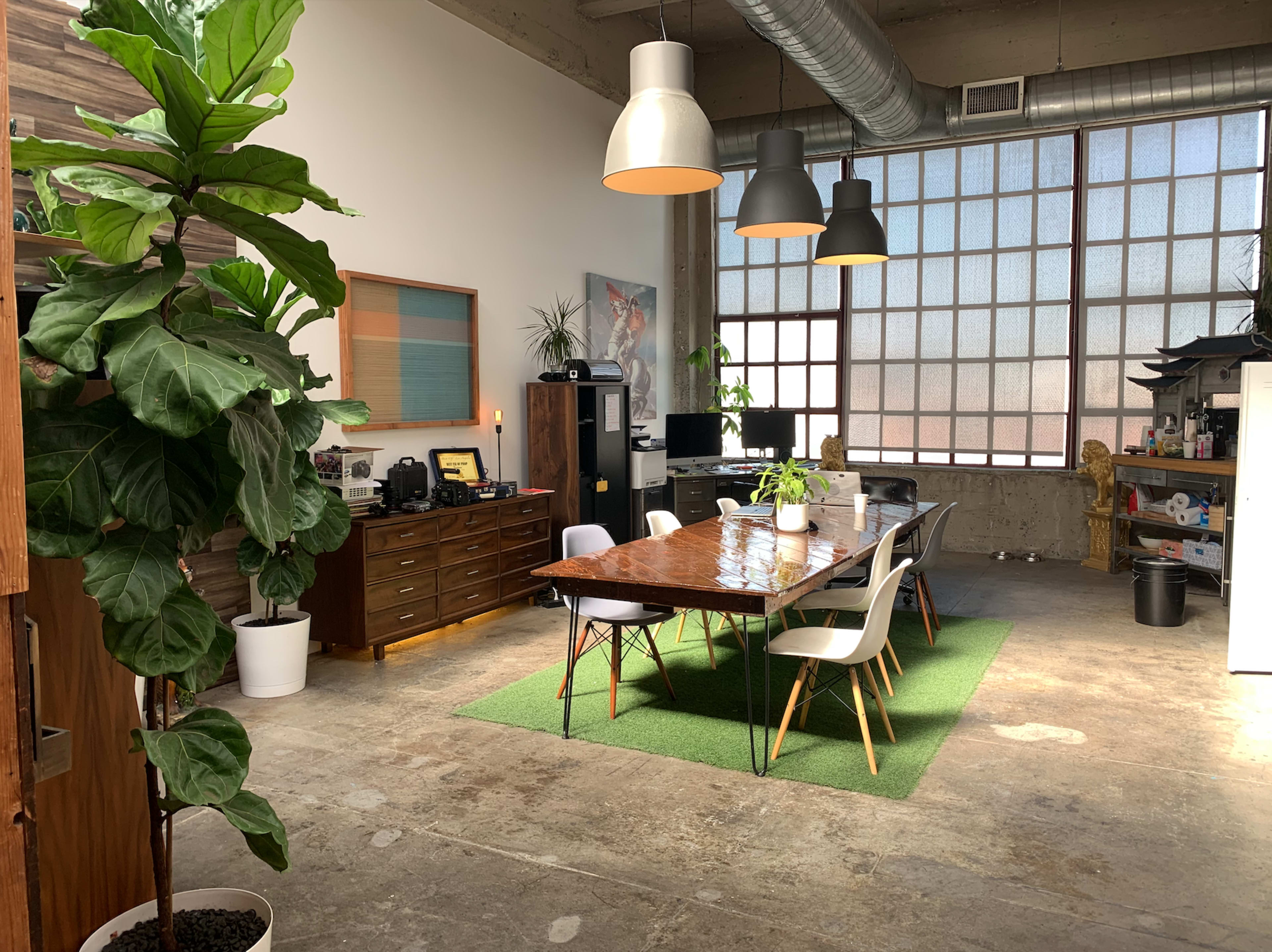 A spacious office interior with a wooden dining table surrounded by white chairs, large windows, and a potted plant in the corner.