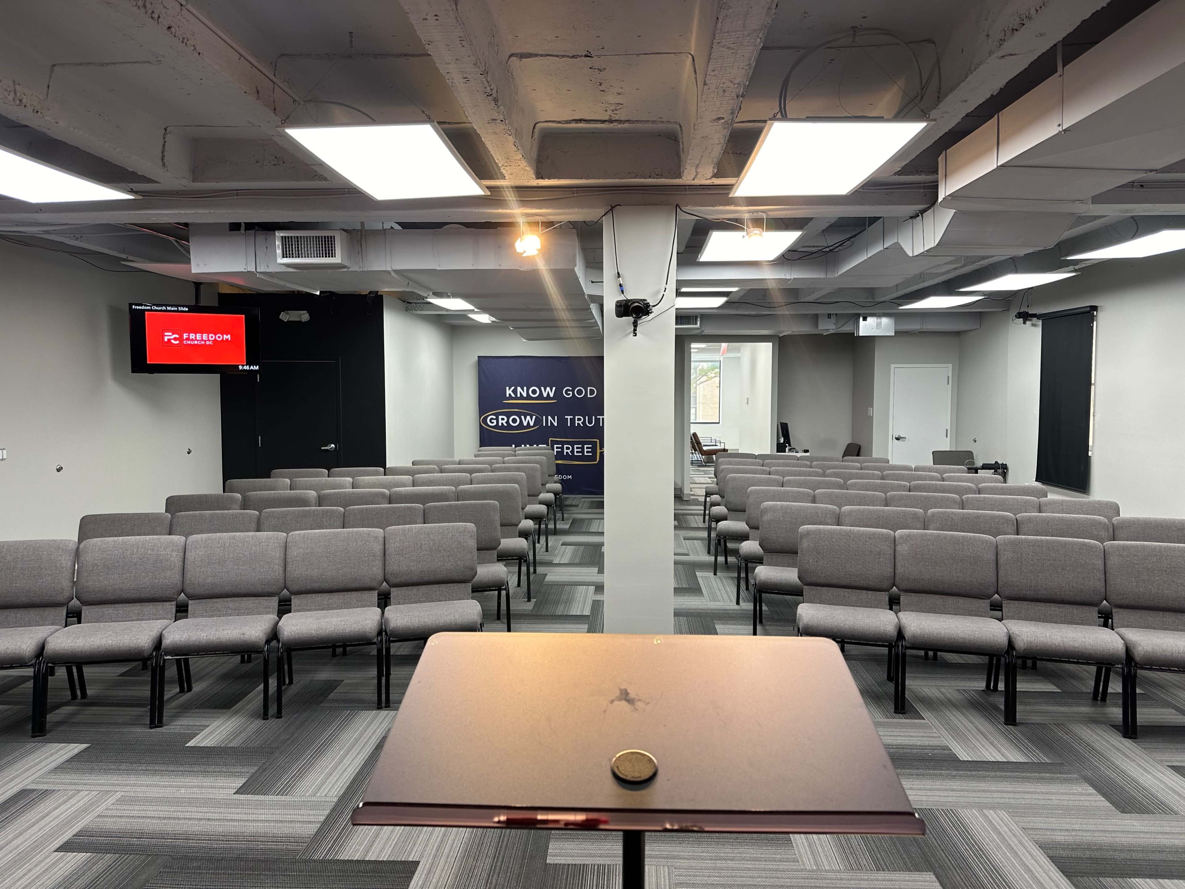 The image shows a spacious room set up for an event, featuring multiple rows of gray chairs facing a podium, with screens and banners on the walls.