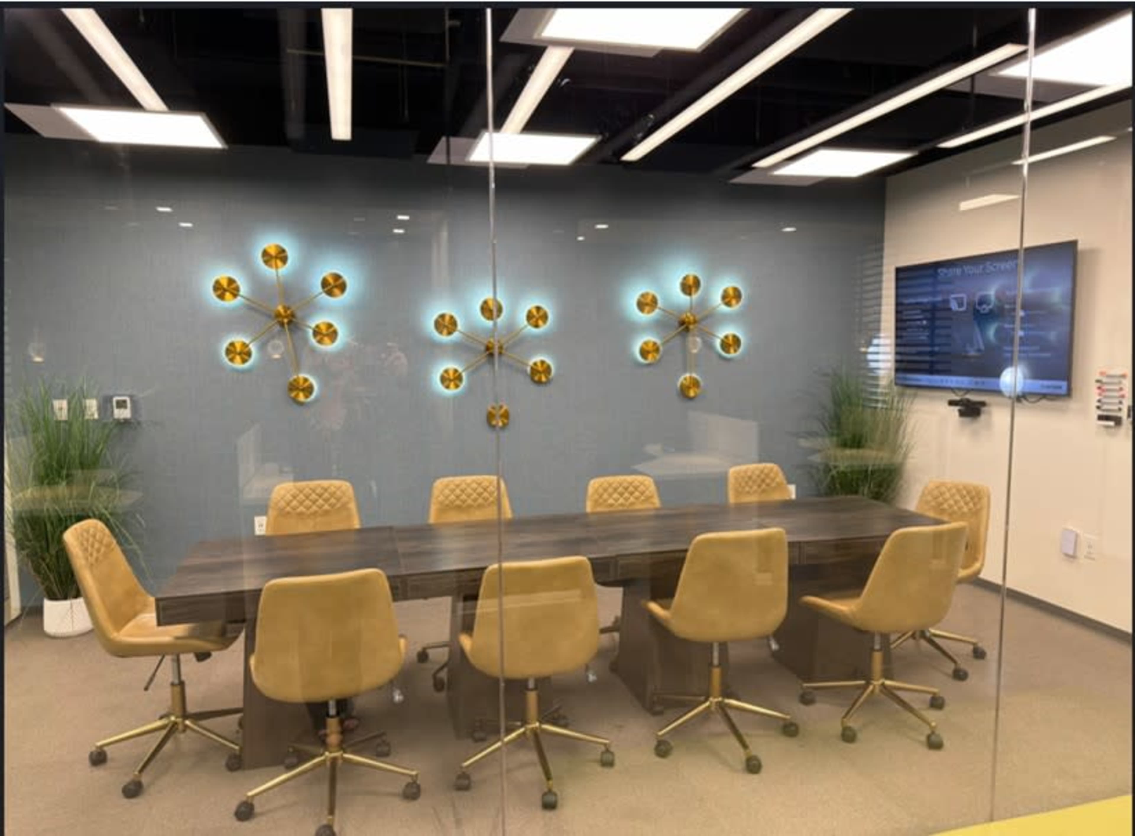 The image shows a modern conference room with a long table surrounded by eight tan chairs, glass walls, and decorative wall accents.