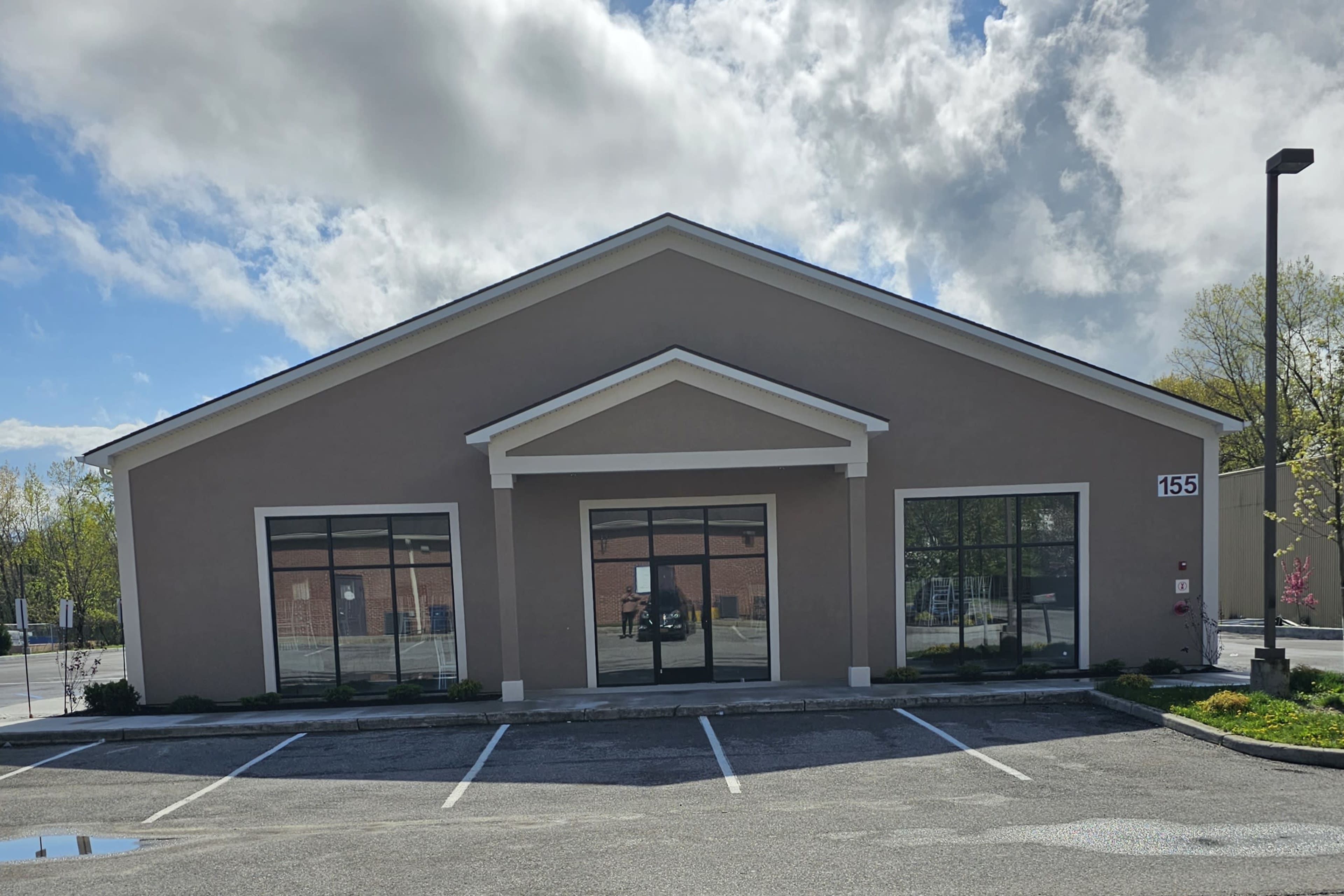 A single-story commercial building with large front windows and a peaked roof, located at 155, sits in a parking lot under a partly cloudy sky.