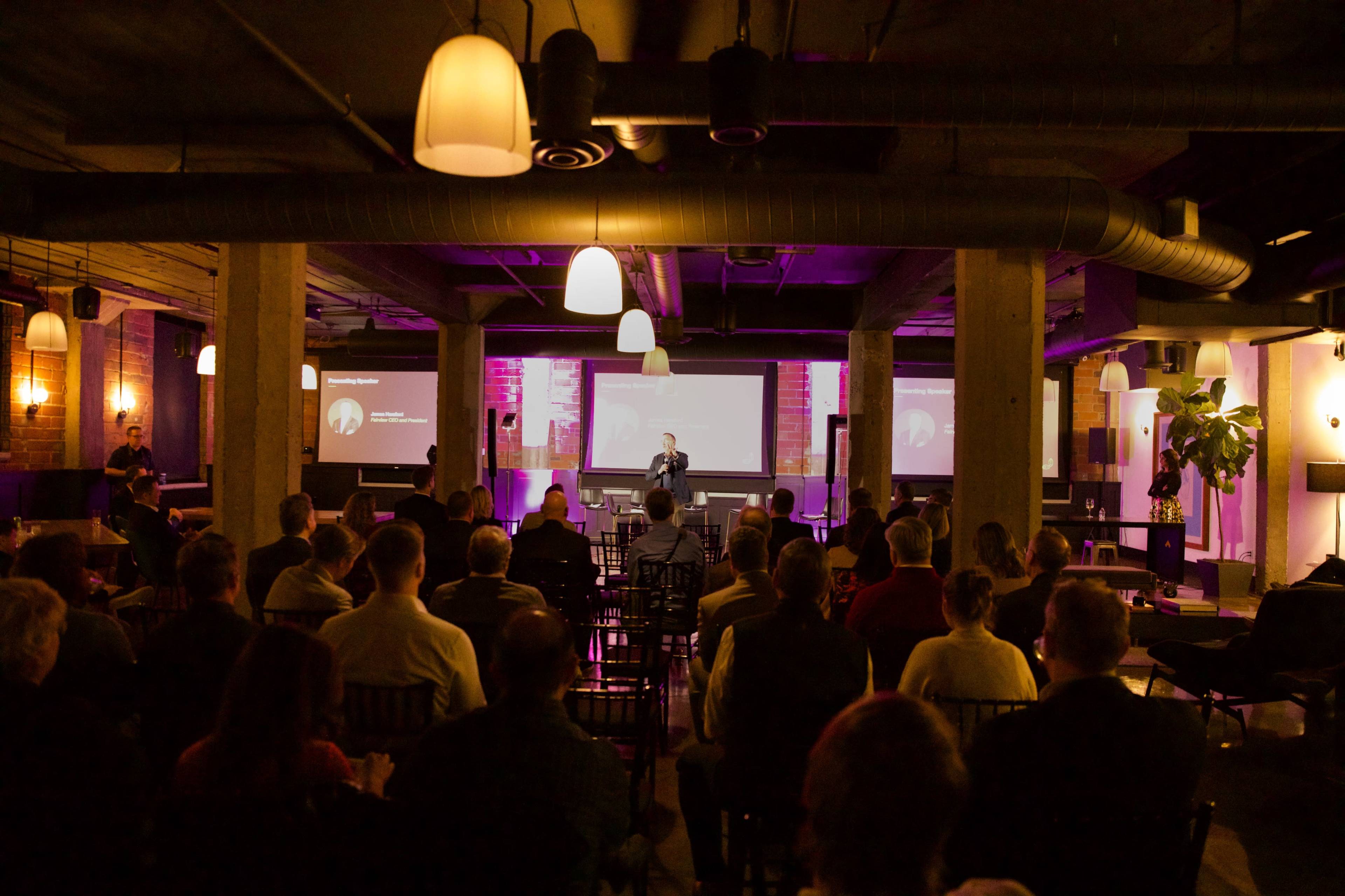 A speaker addresses an audience in a dimly lit venue featuring multiple screens and modern lighting.