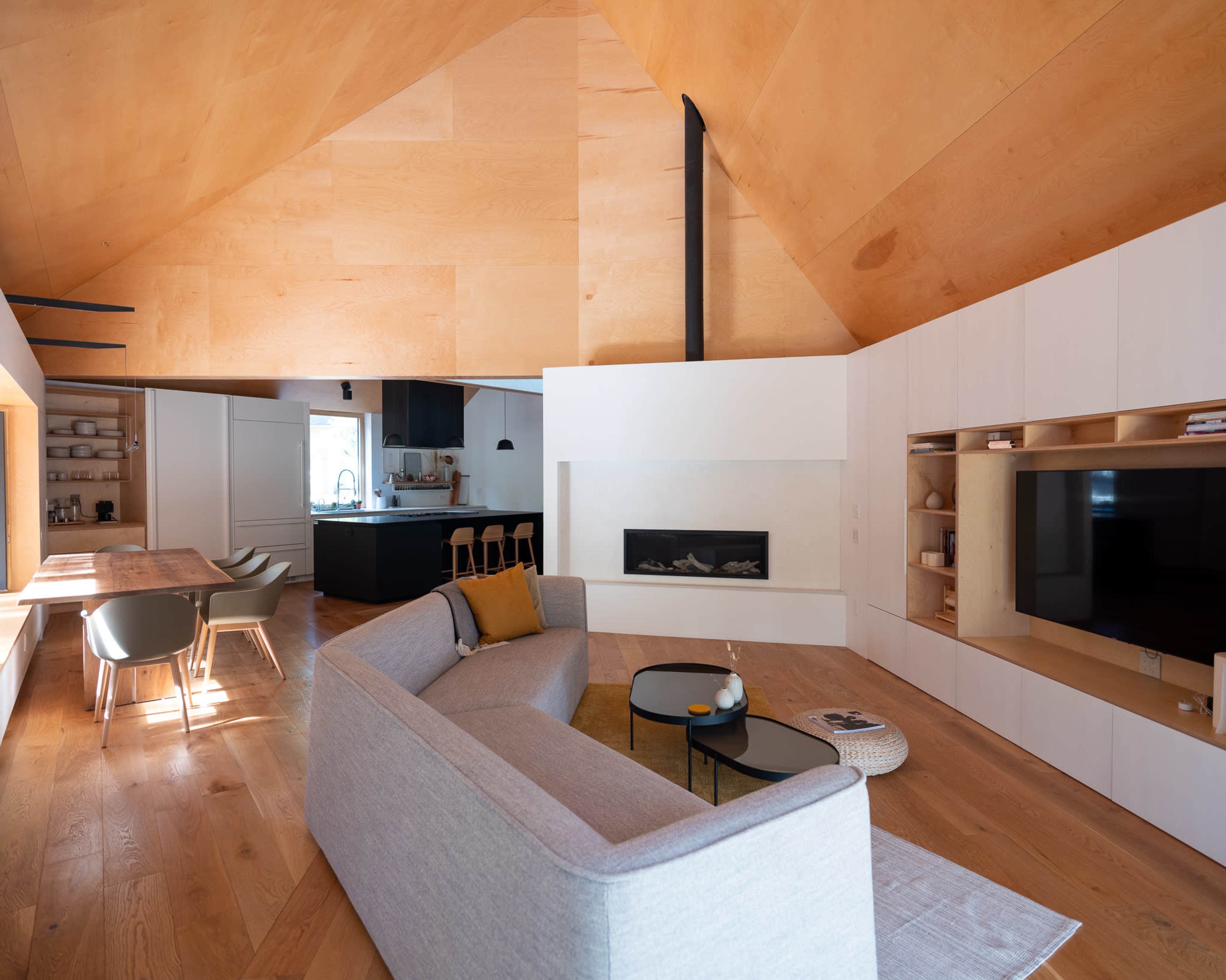 The image shows a modern, open-concept living room with a large sofa, a dining area, and a sleek kitchen, all featuring wooden accents and a minimalist design.