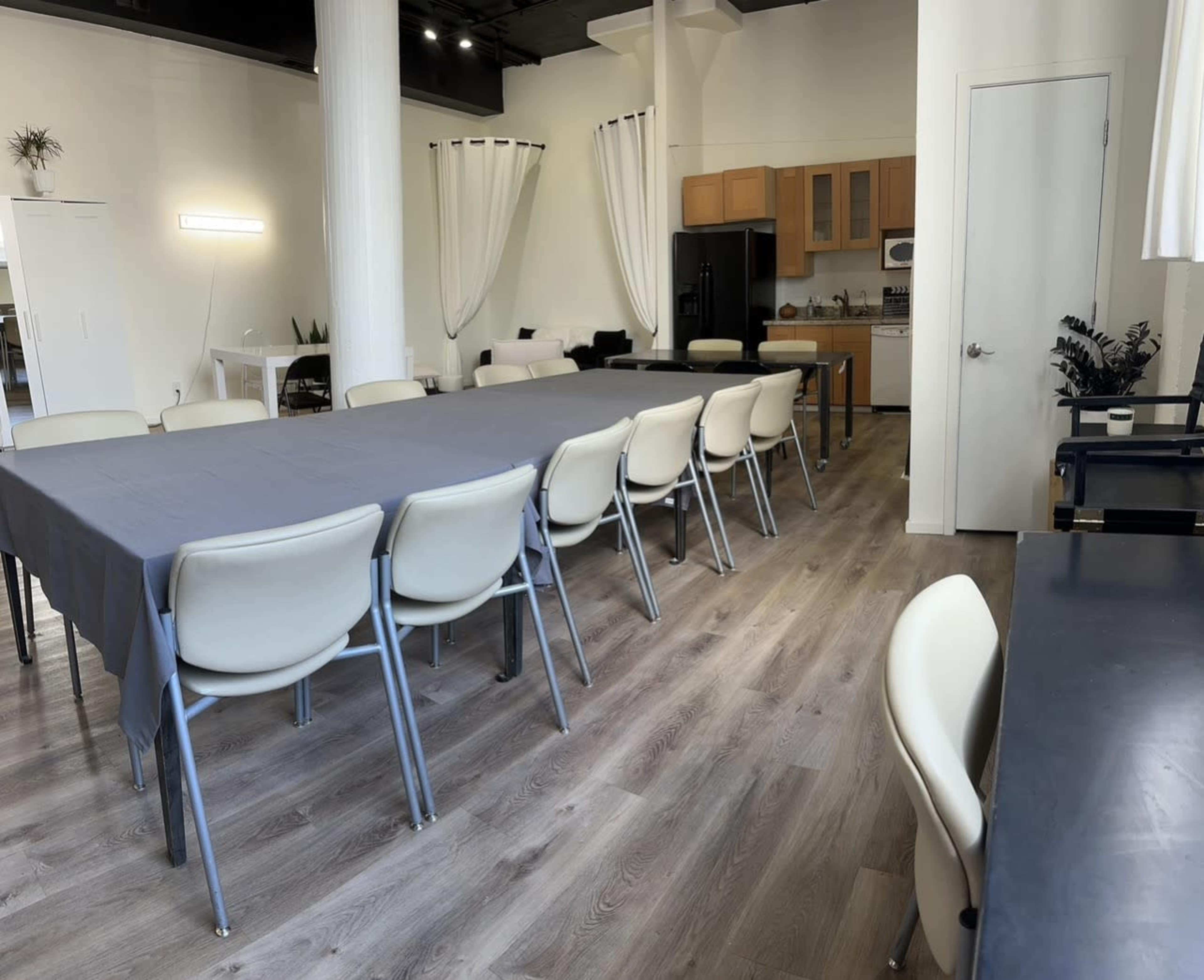 The image shows a spacious room with a long table set with chairs, and a kitchenette visible in the background.