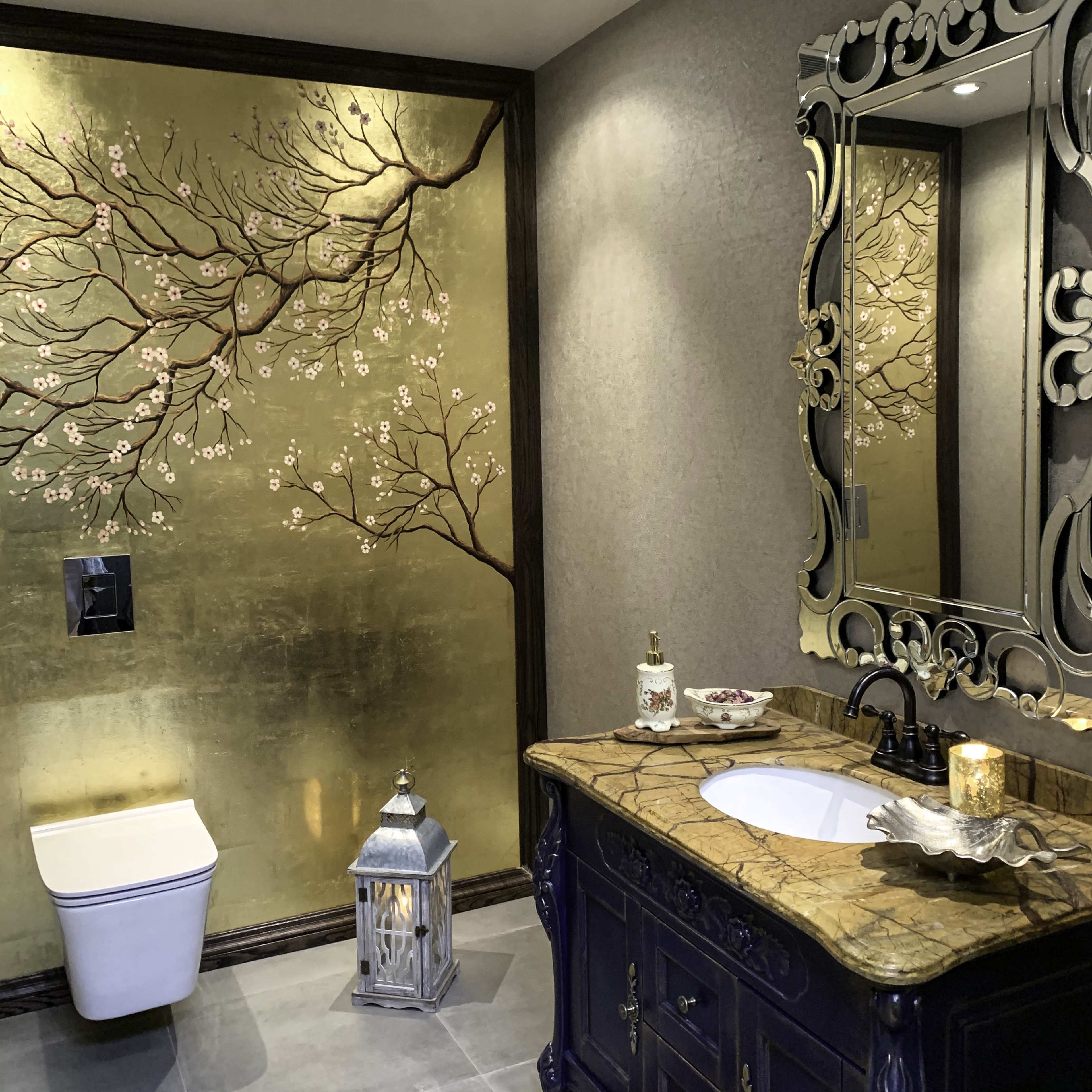 A bathroom features a gold-painted wall with a floral design, a dark wooden vanity with a marble countertop, a modern toilet, and decorative elements including a lantern and a mirror.