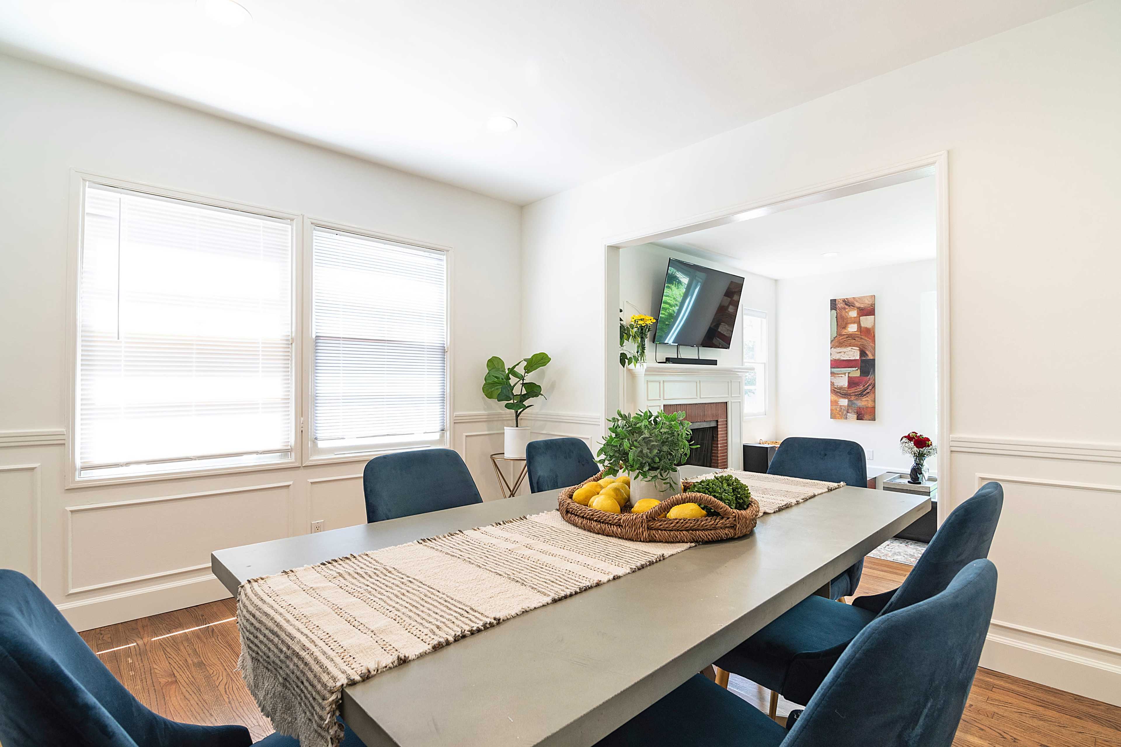 A dining room features a rectangular table with a central arrangement of fruits and greenery, surrounded by blue upholstered chairs, and natural light coming through multiple windows.