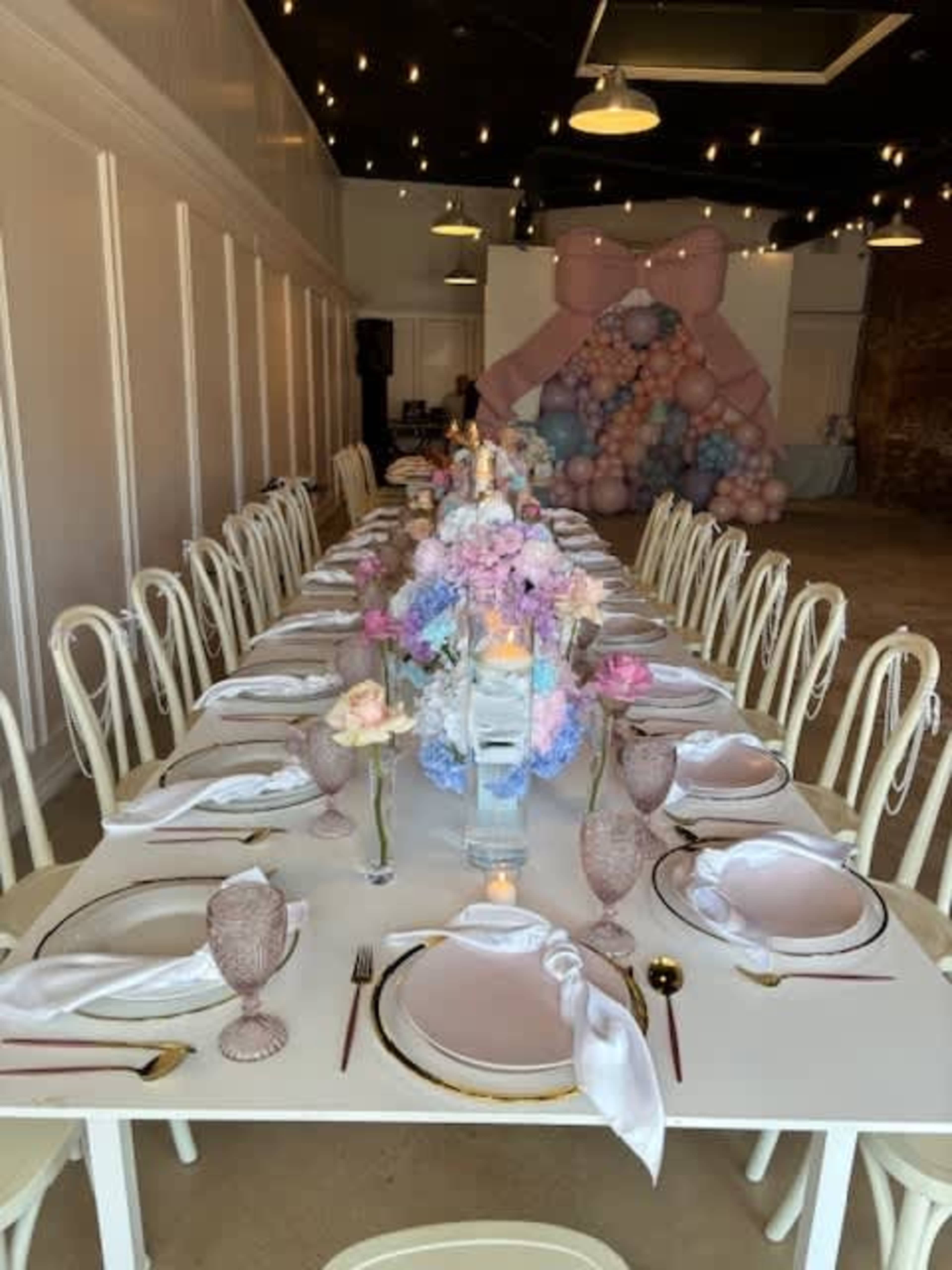 A long, elegantly set table with pink and pastel floral arrangements, glassware, and white napkins is arranged in a venue with decorative balloon installations in the background.