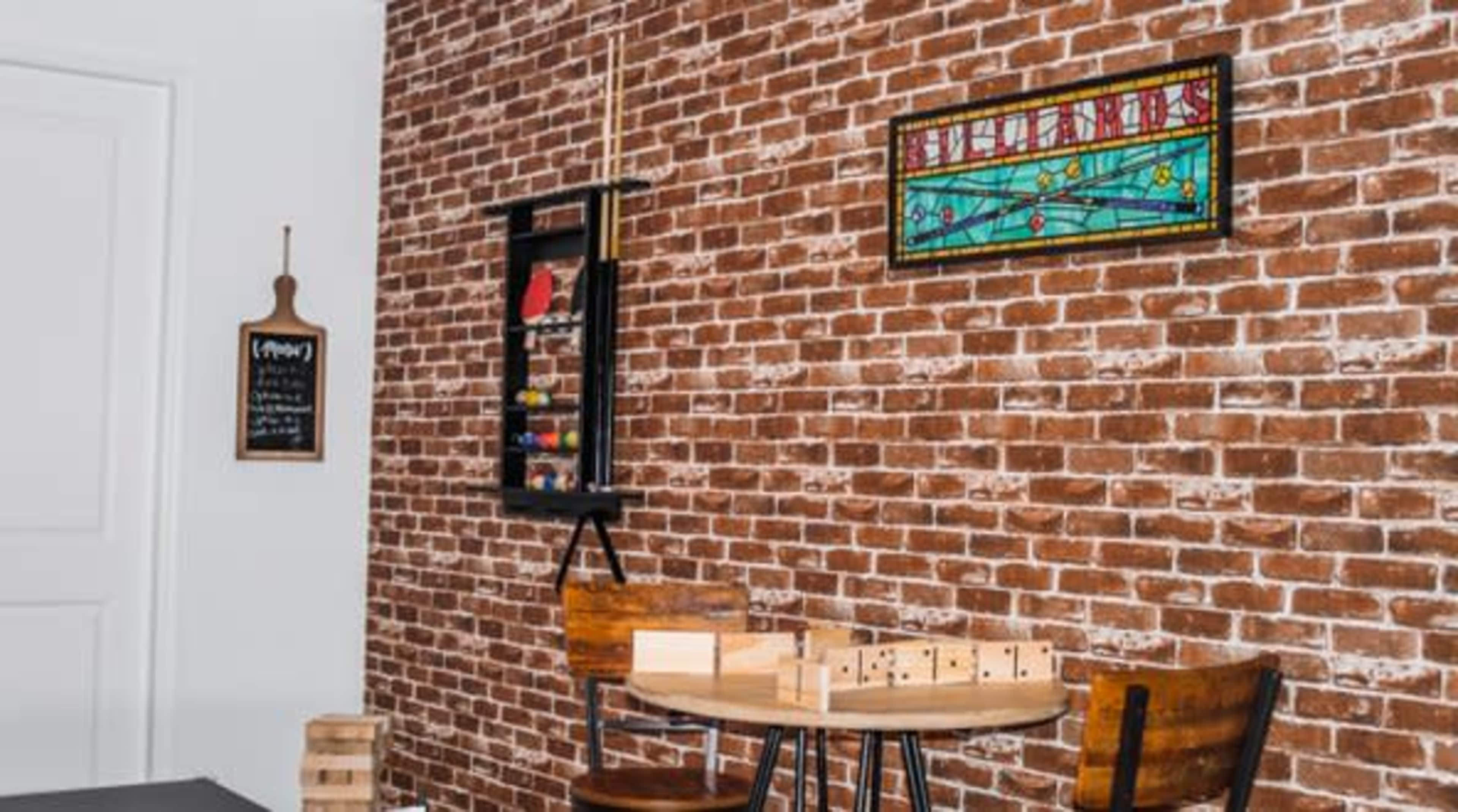 A cozy room features a brick wall with a stained glass window, a round table with wooden chairs, and various game pieces displayed on a shelf.