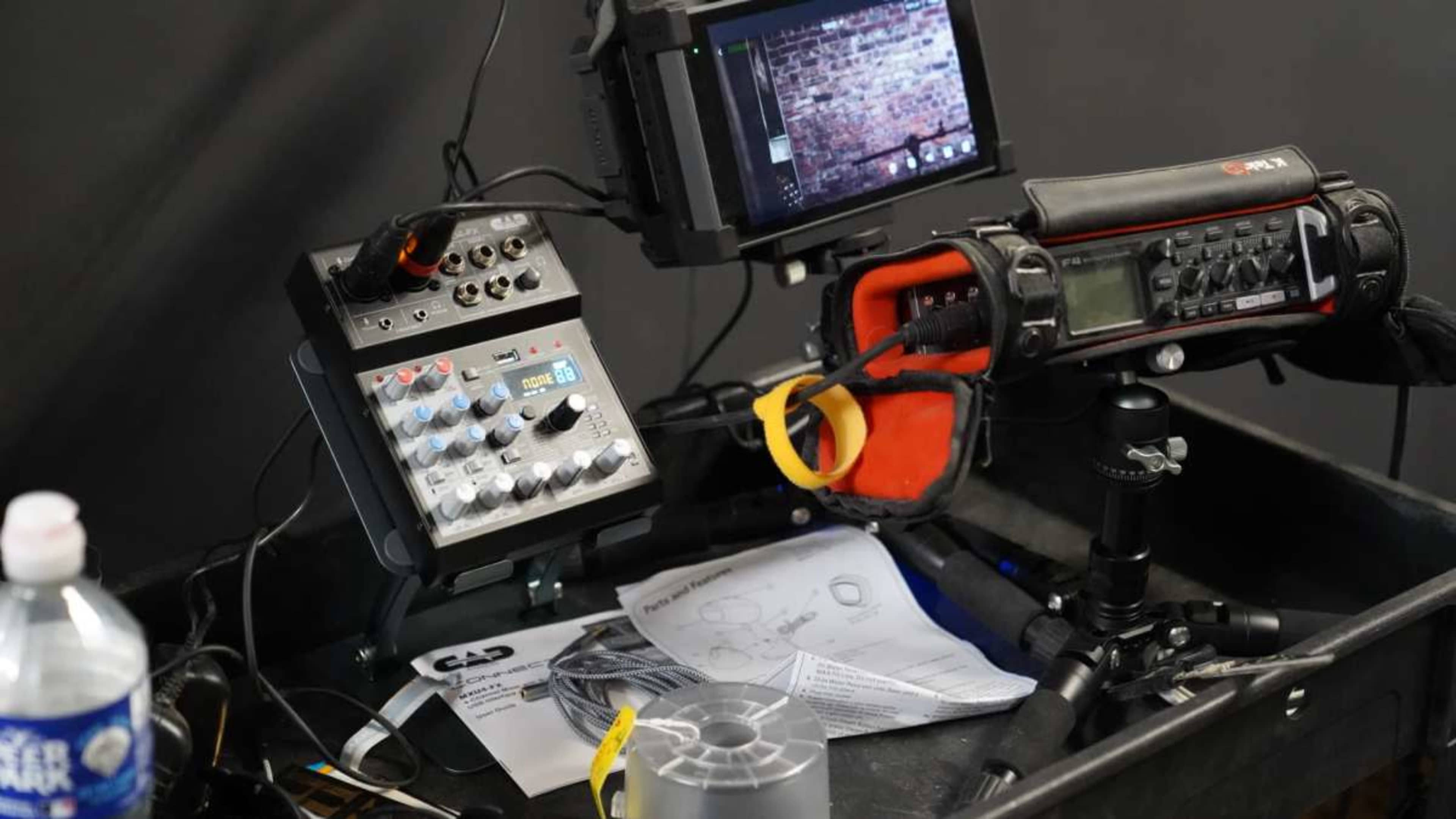 The image shows a sound mixer, a recording device, and a monitor arranged on a black cart, alongside a water bottle and documentation.