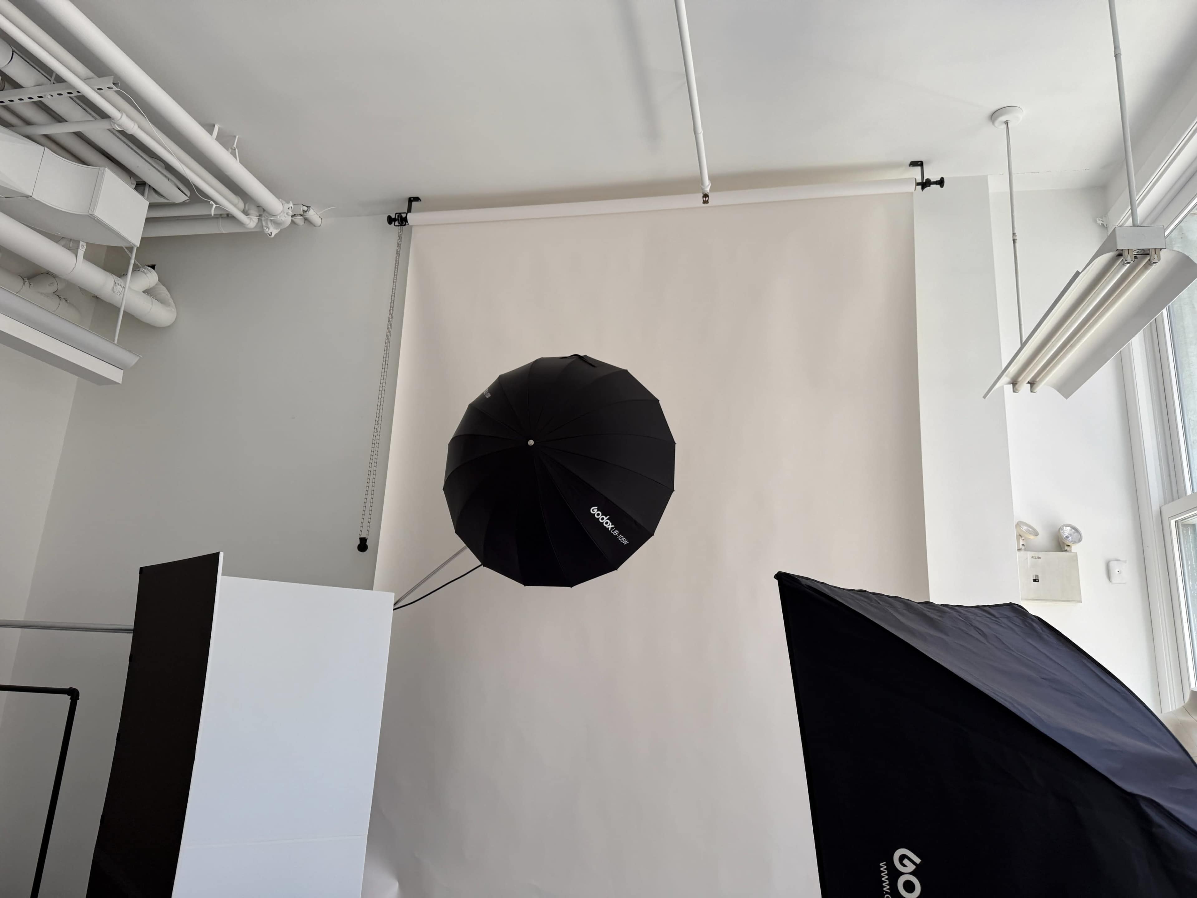 The image shows a black umbrella suspended from the ceiling in a photo studio with a plain white backdrop and softboxes arranged on the floor.