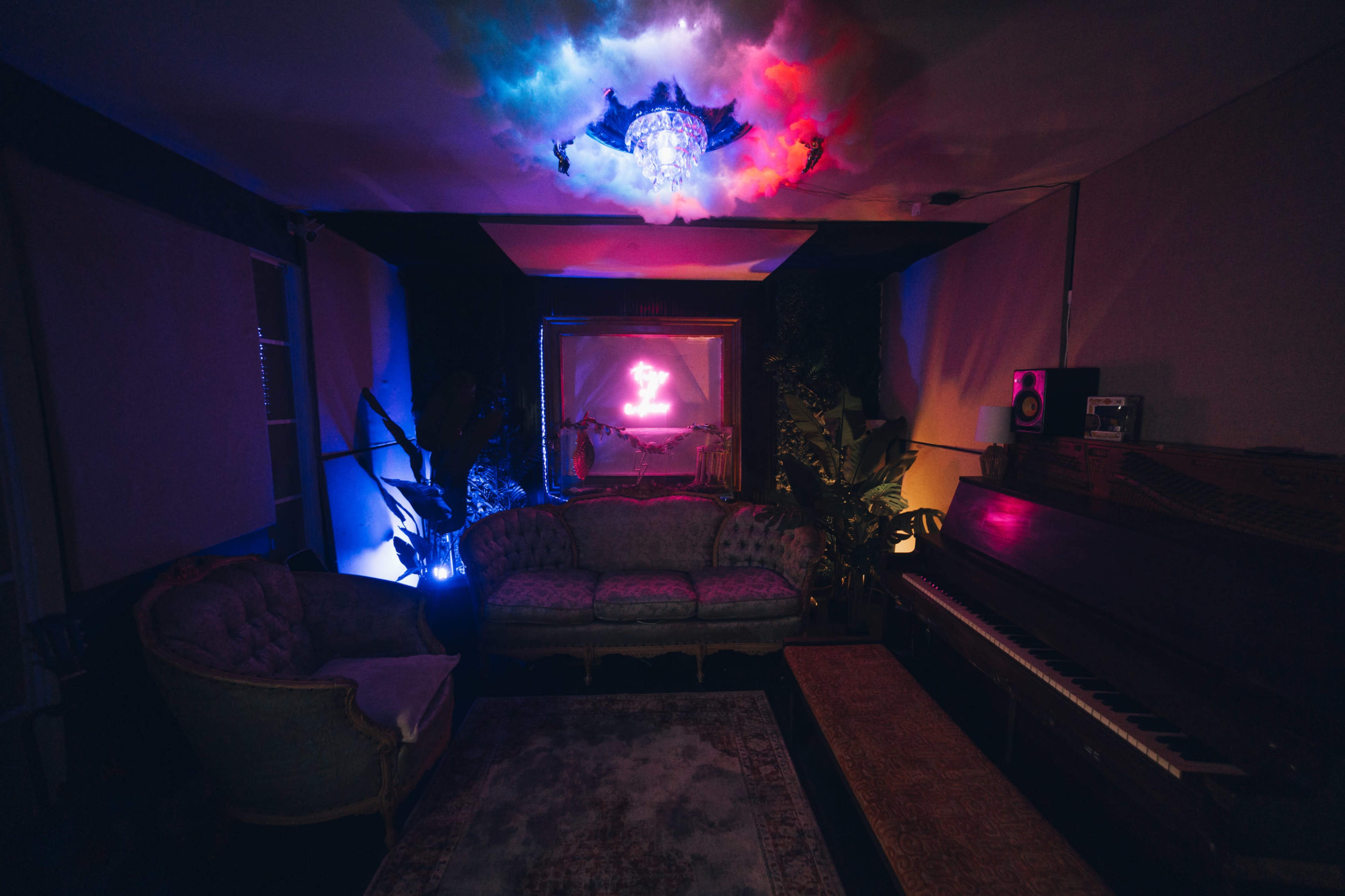 The image shows a dimly lit room with vintage furniture, including a sofa and an armchair, a piano, and colorful lighting illuminating the space.