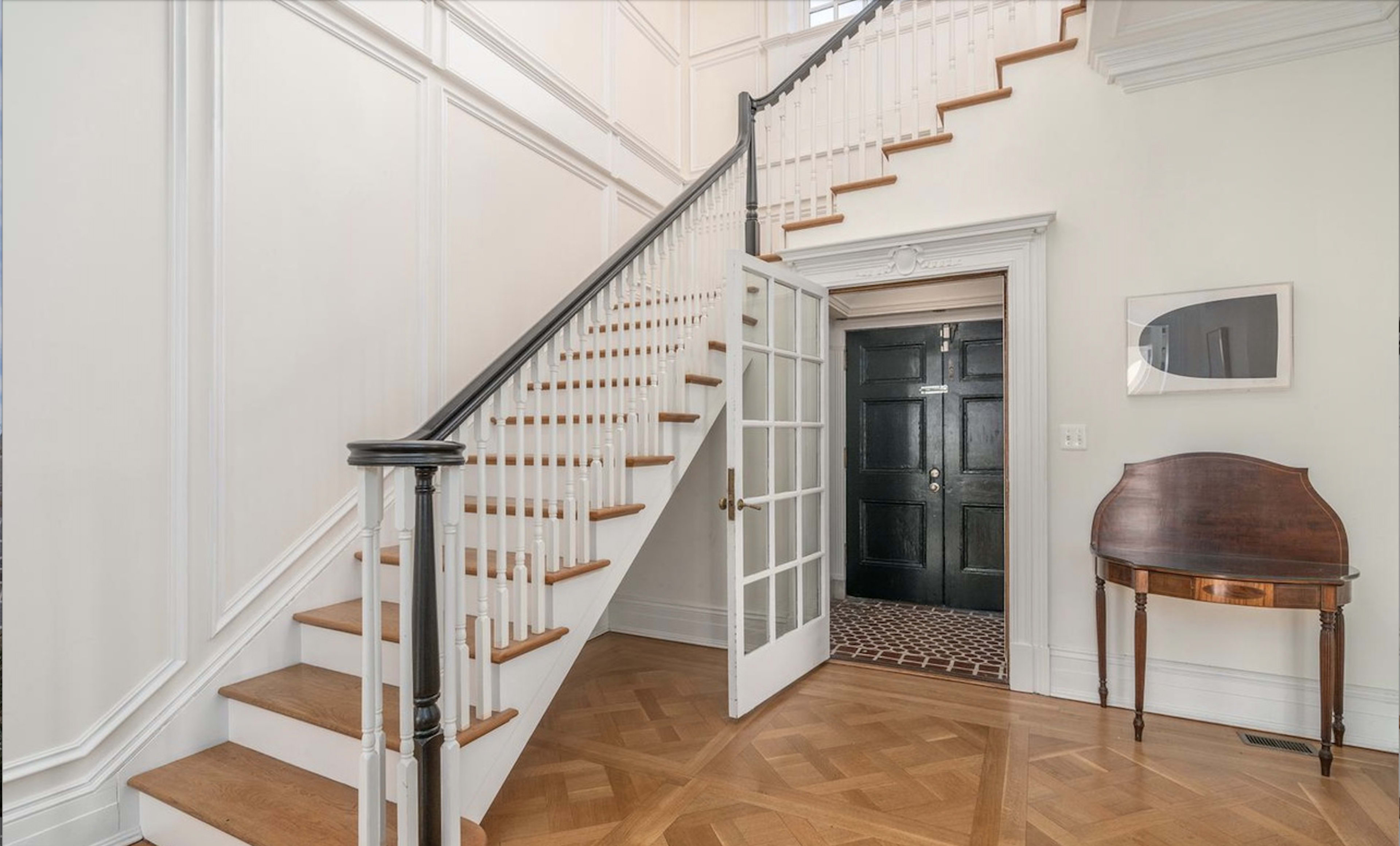 A spacious entryway featuring a staircase with wooden steps and a white railing, leading to an upper level, alongside a dark front door and a small wooden table against the wall.