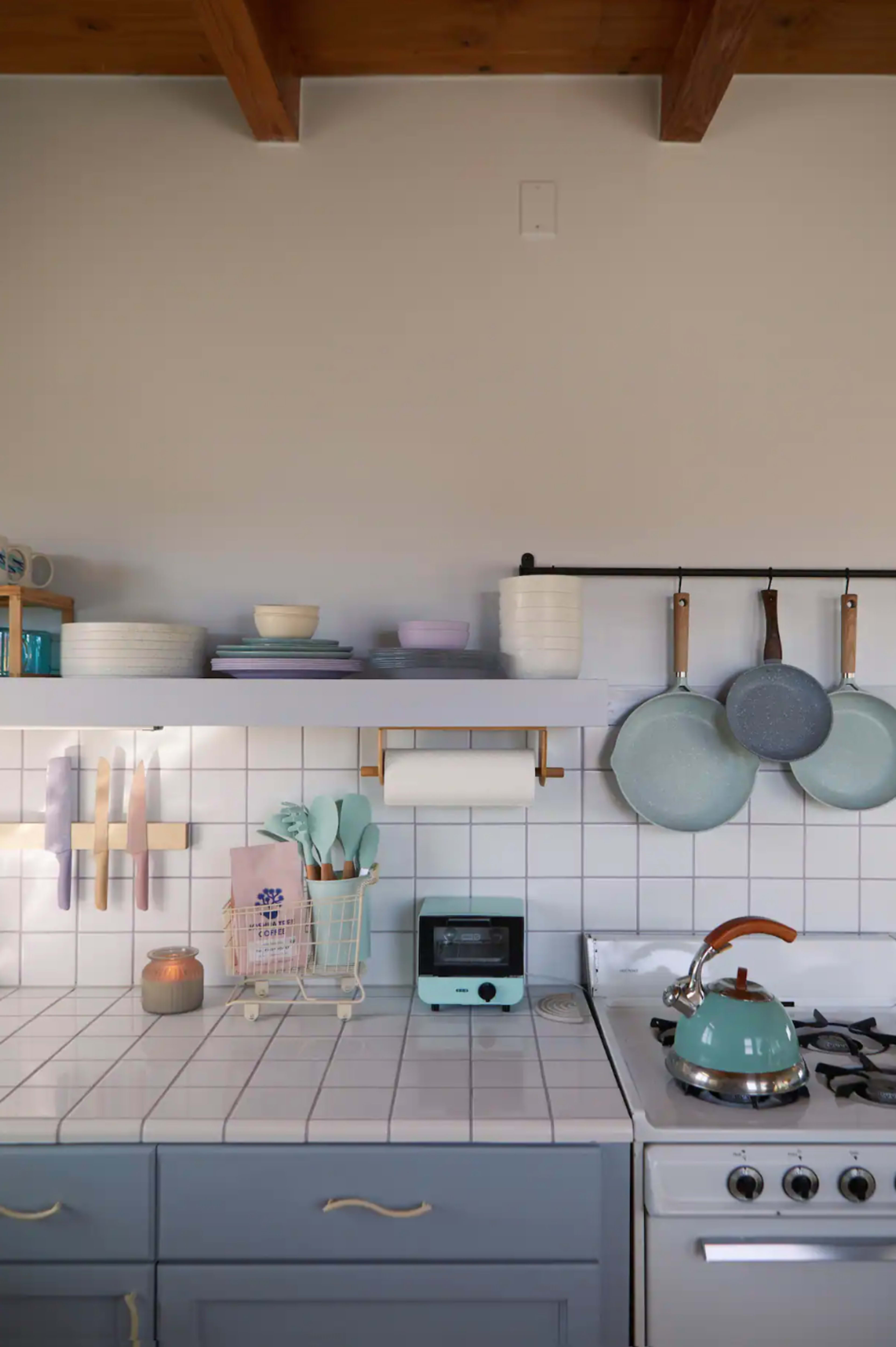 A bright kitchen features a tiled countertop with utensils and bowls, a blue kettle on the stove, and hanging pots above.
