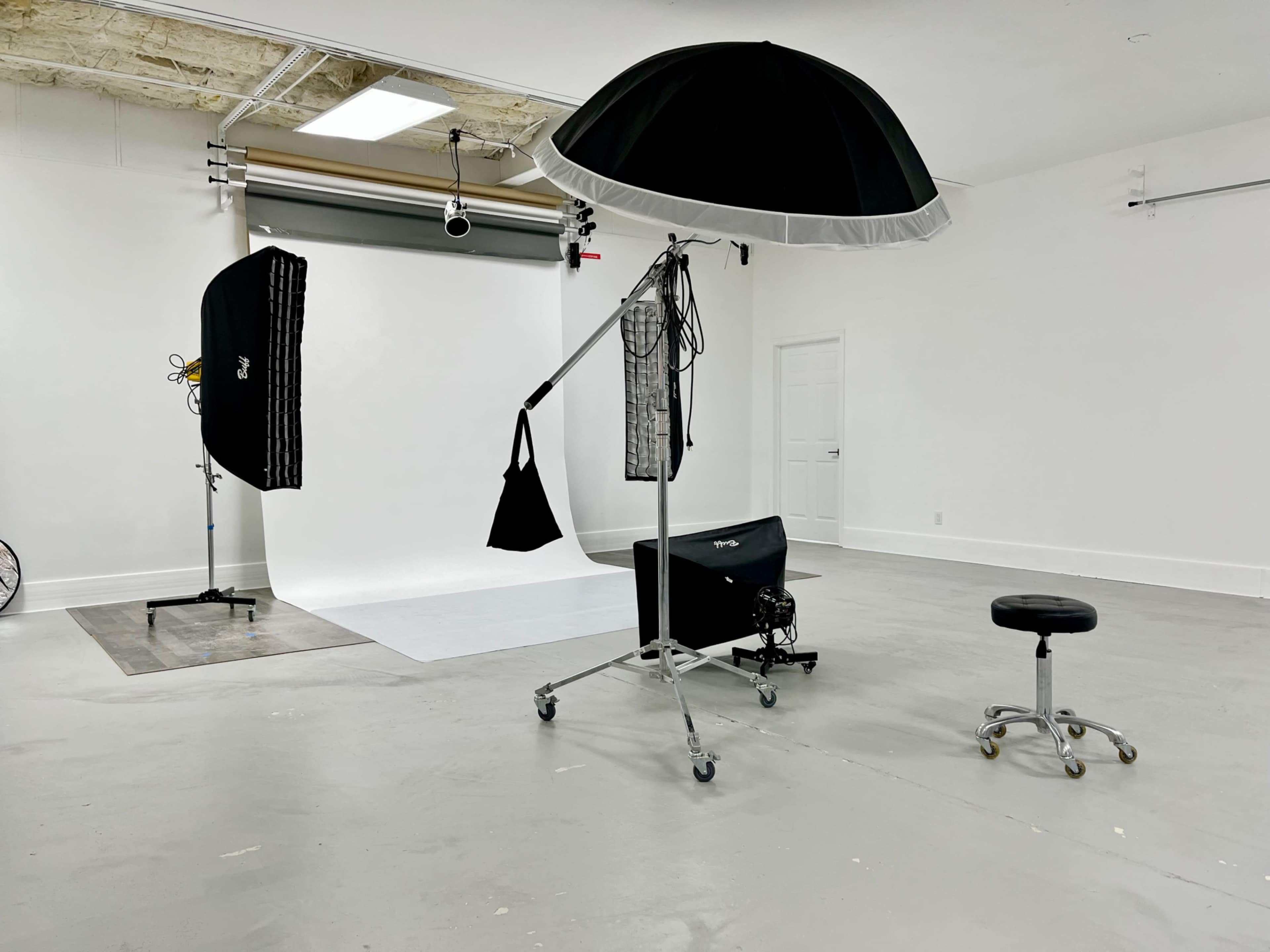 The image shows a photography studio with a backdrop, lighting equipment, a stand for reflectors, and a rolling stool on a polished concrete floor.