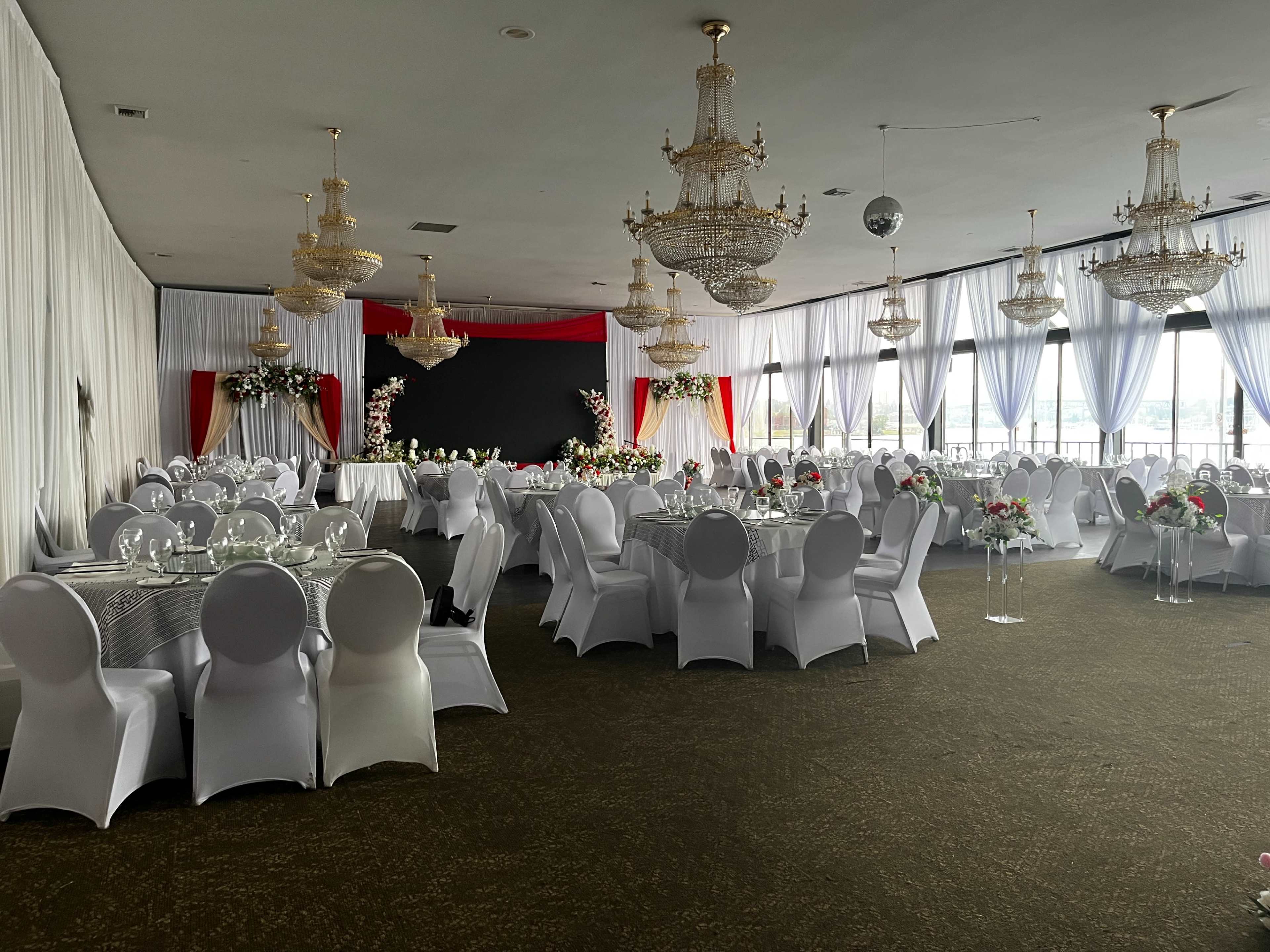 The image shows an elegantly arranged banquet hall with round tables covered in white linens and decorated with floral centerpieces, chandeliers hanging from the ceiling, and large windows allowing natural light.