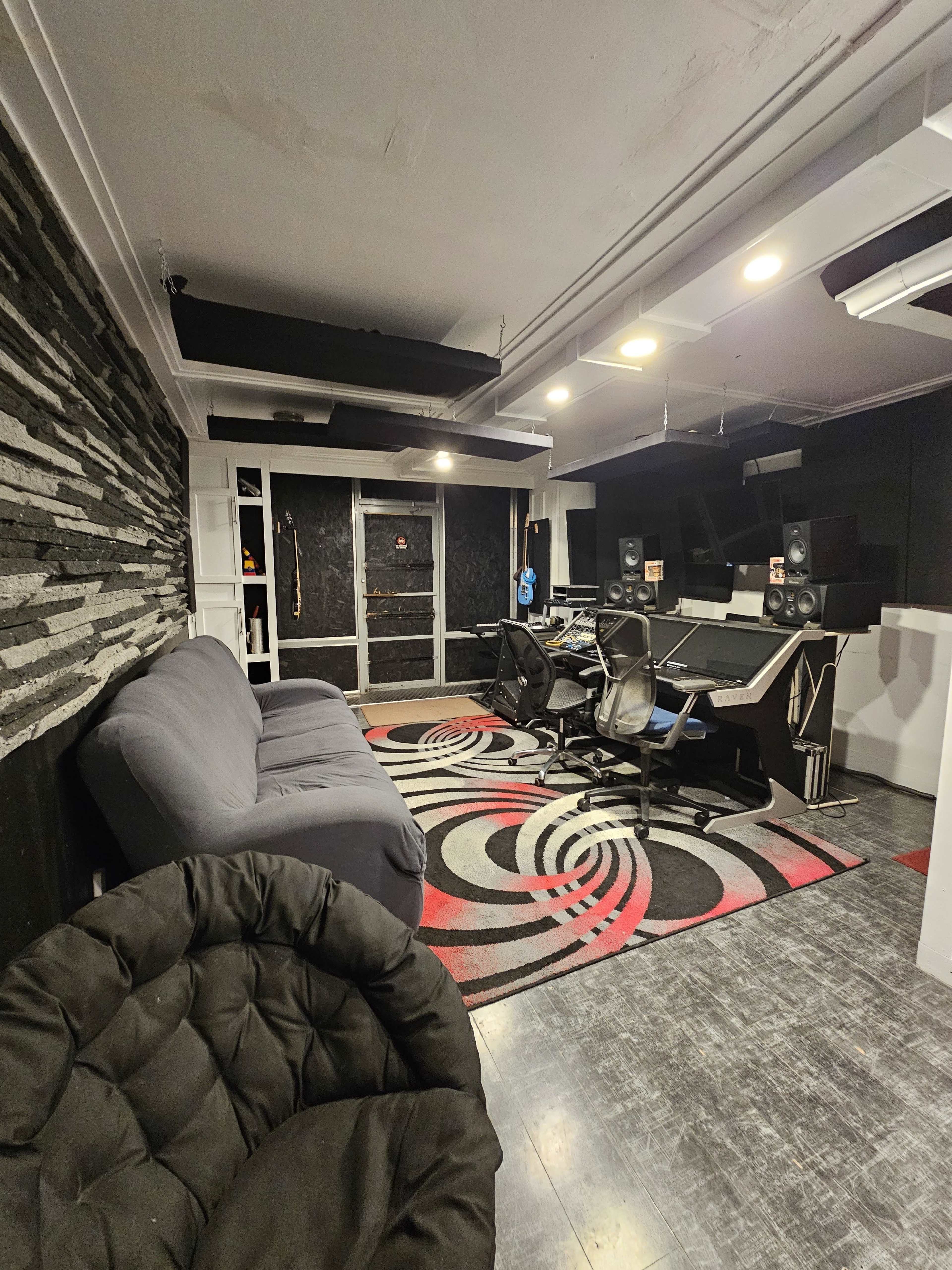 The image features a modern recording studio with a black and white patterned rug, multiple workstations, soundproofing panels on the walls, and a couch in the corner.