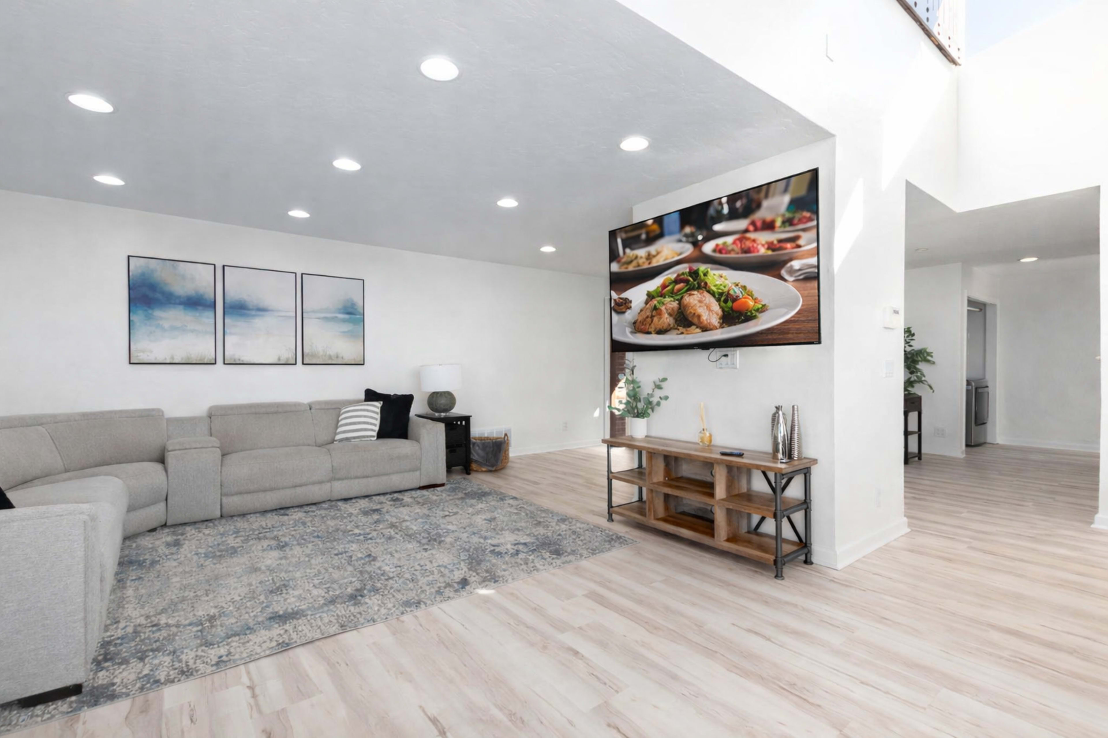 A modern living room features a gray sectional sofa, a wooden TV stand, and a large wall-mounted television displaying food, with three framed artworks on the wall.