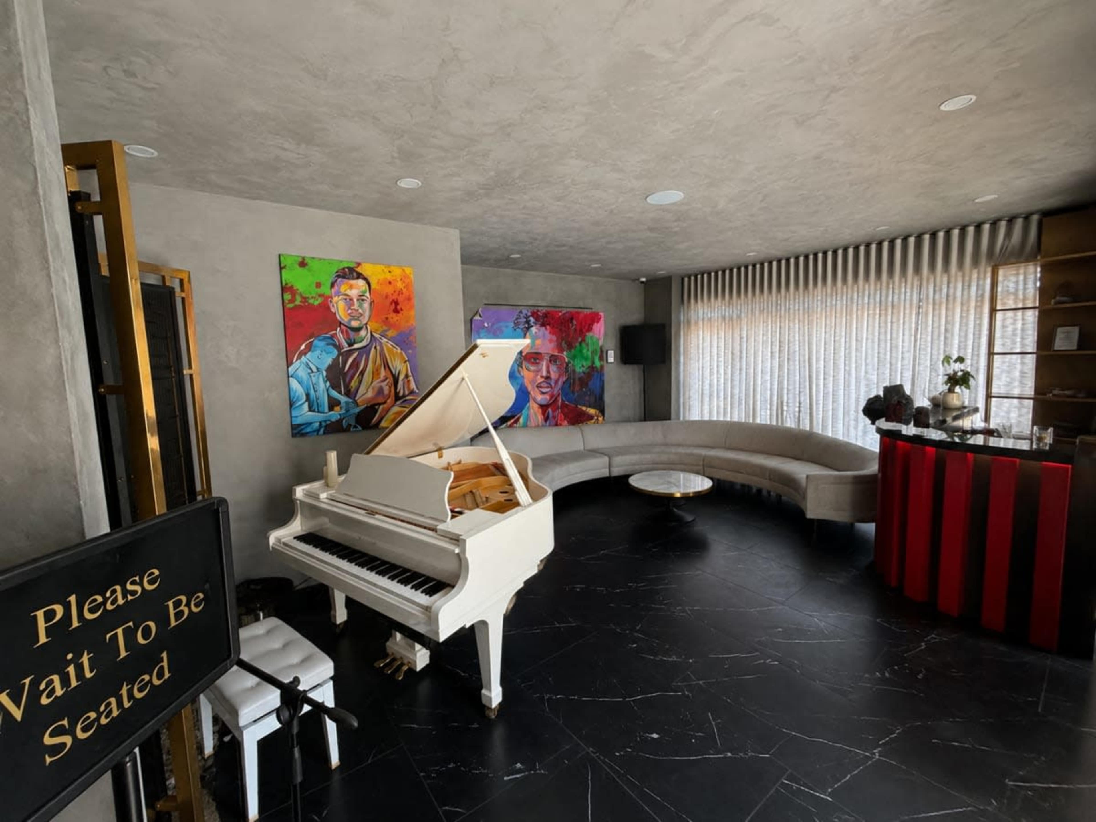 The image shows a modern lounge area featuring a white grand piano, two colorful paintings on the wall, a curved gray sofa, a bar cart, and a "Please Wait To Be Seated" sign.