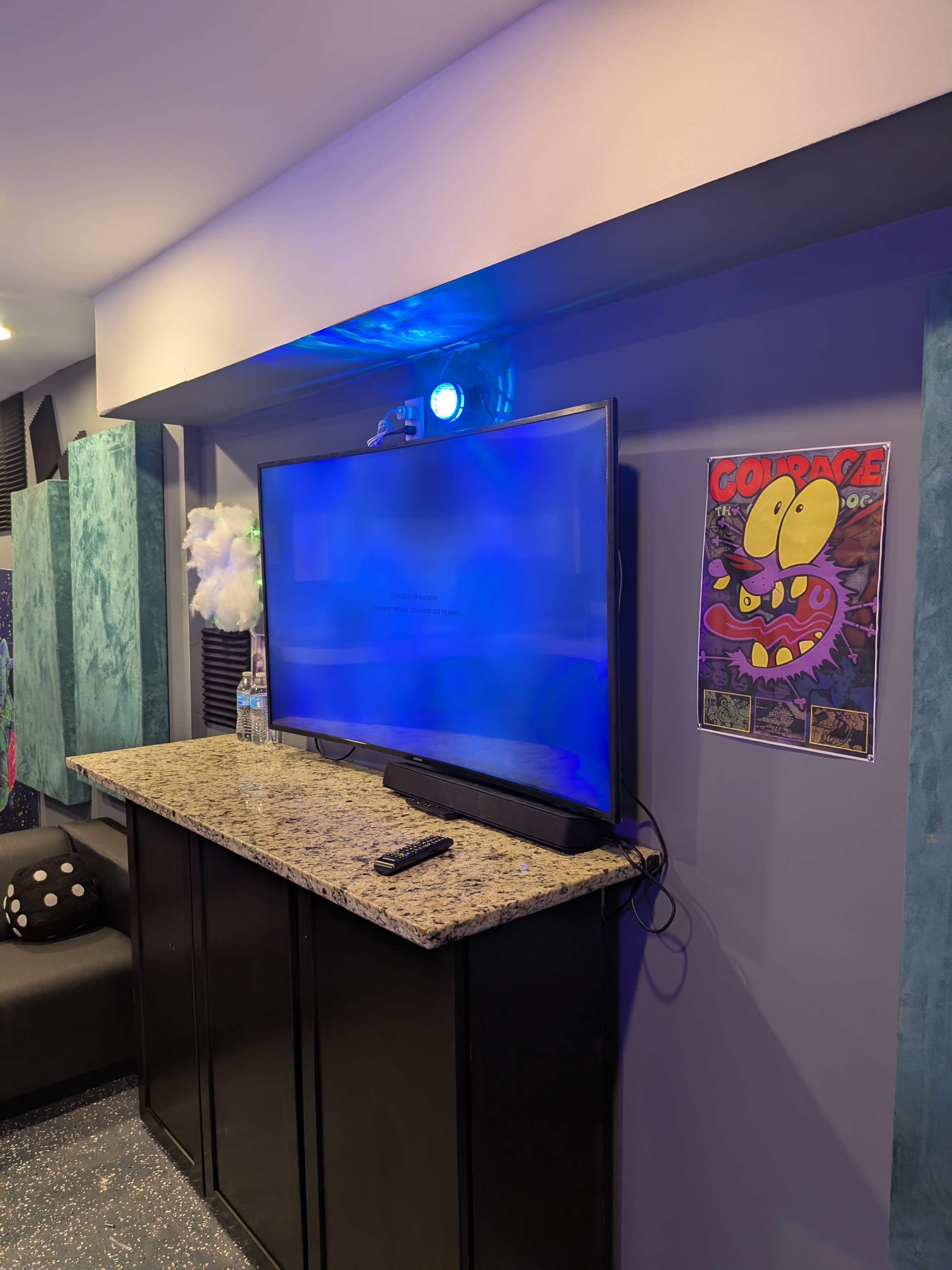 The image shows a modern entertainment setup featuring a large flat-screen TV mounted on a wall above a granite countertop, with a colorful poster and a remote control placed on the surface below.