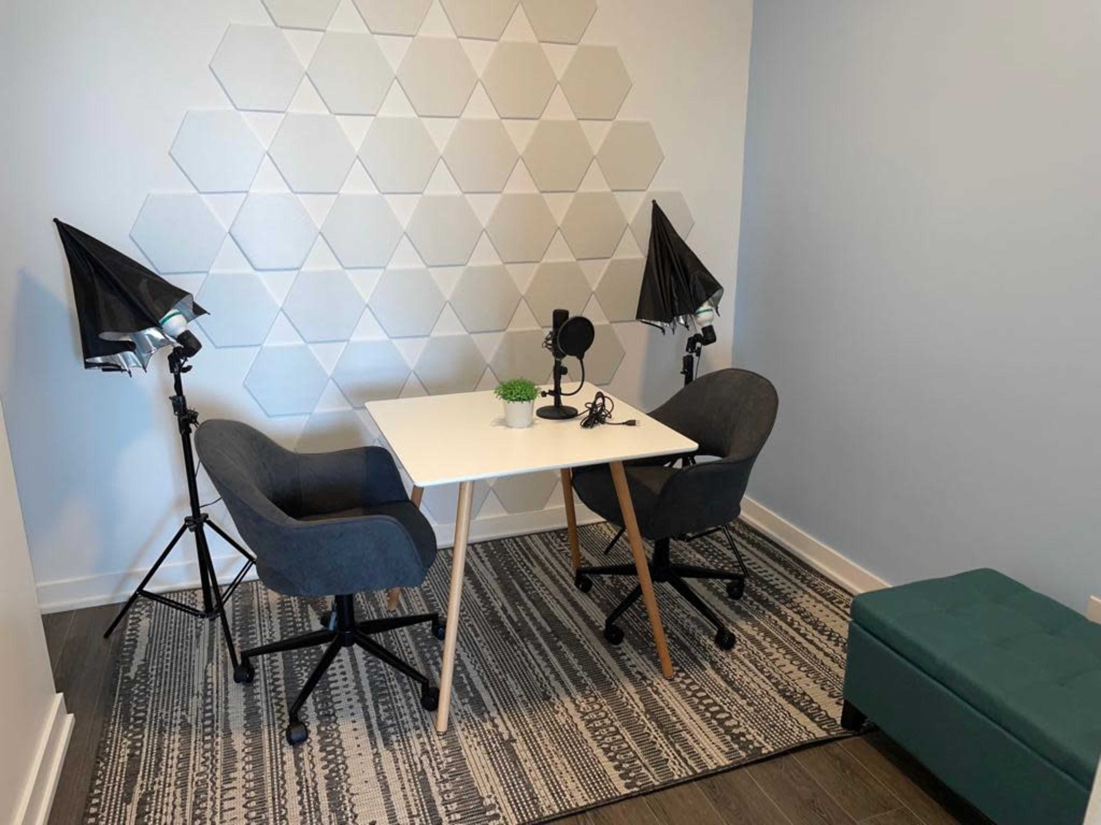 A small, modern room features a white table with two chairs, a microphone setup, and lighting equipment against a geometric patterned wall.