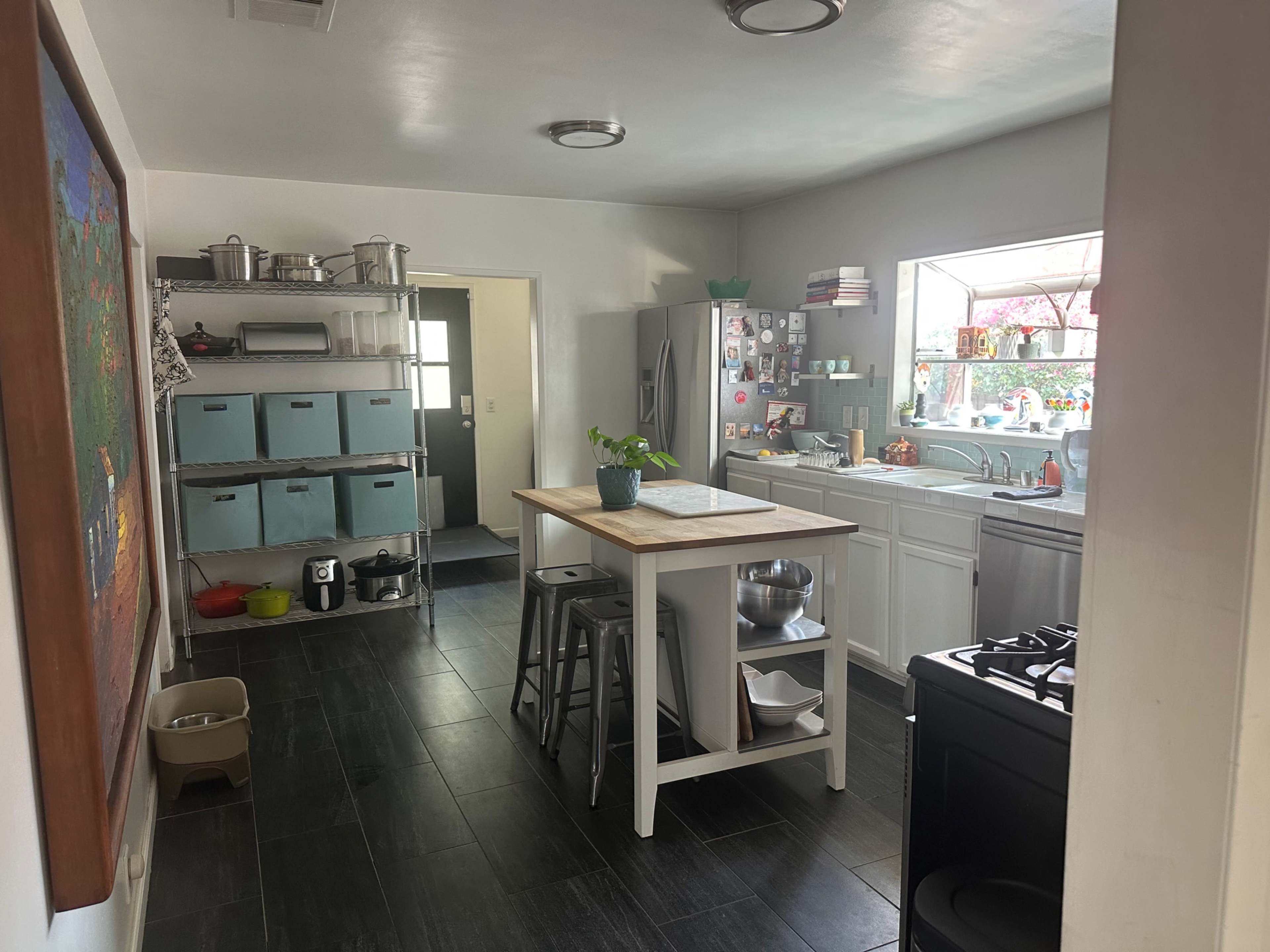 The image shows a modern kitchen with white cabinetry, a central island, stainless steel appliances, and large windows allowing natural light.