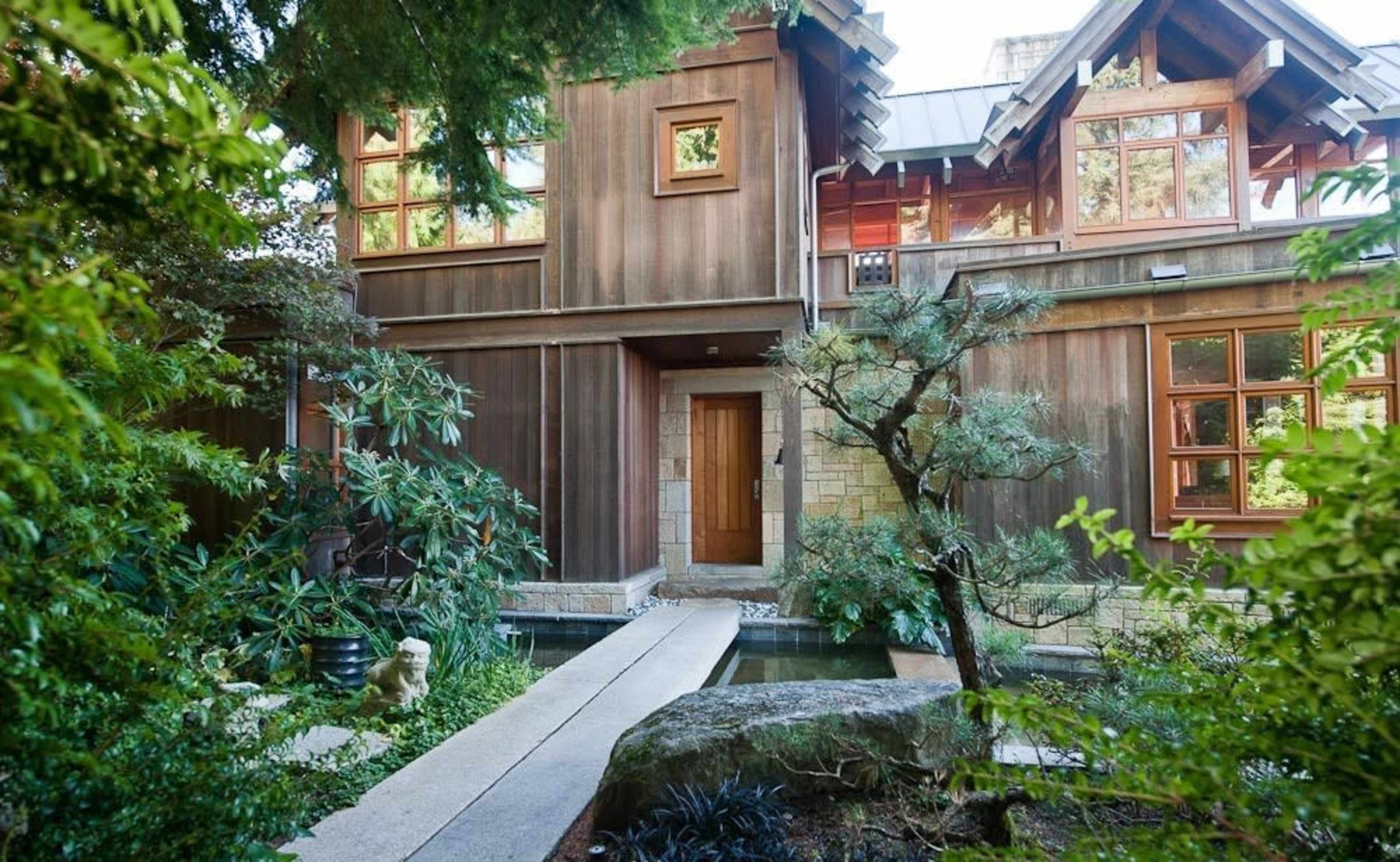 The image shows a wooden house surrounded by lush greenery, with a stone pathway leading to the entrance.