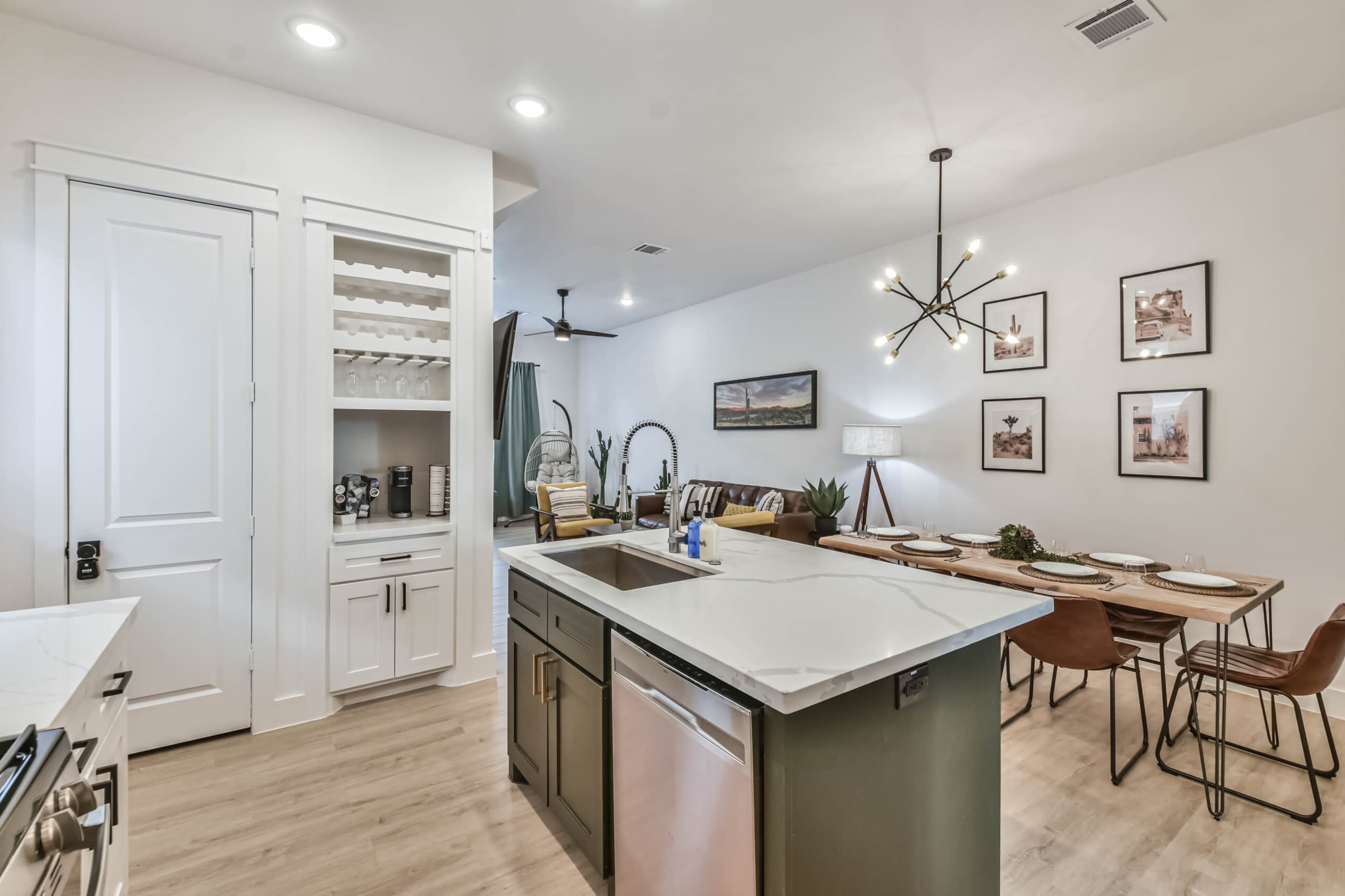 A modern kitchen with a central island, an adjacent dining area featuring a long table and chairs, and a shelf containing kitchenware.