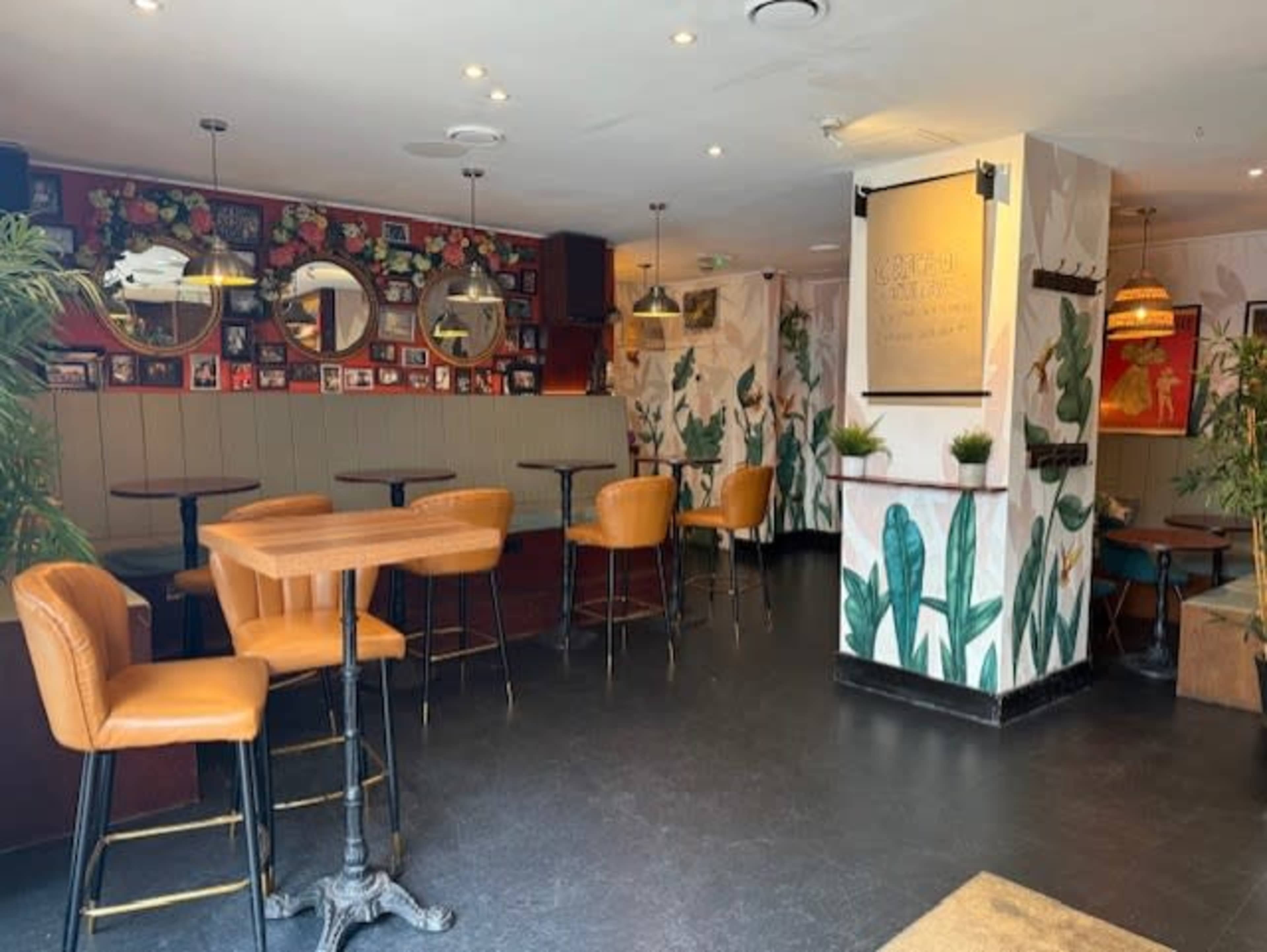 The image shows a cozy bar area featuring high wooden tables with orange leather stools, decorative plants, and framed pictures on the walls.