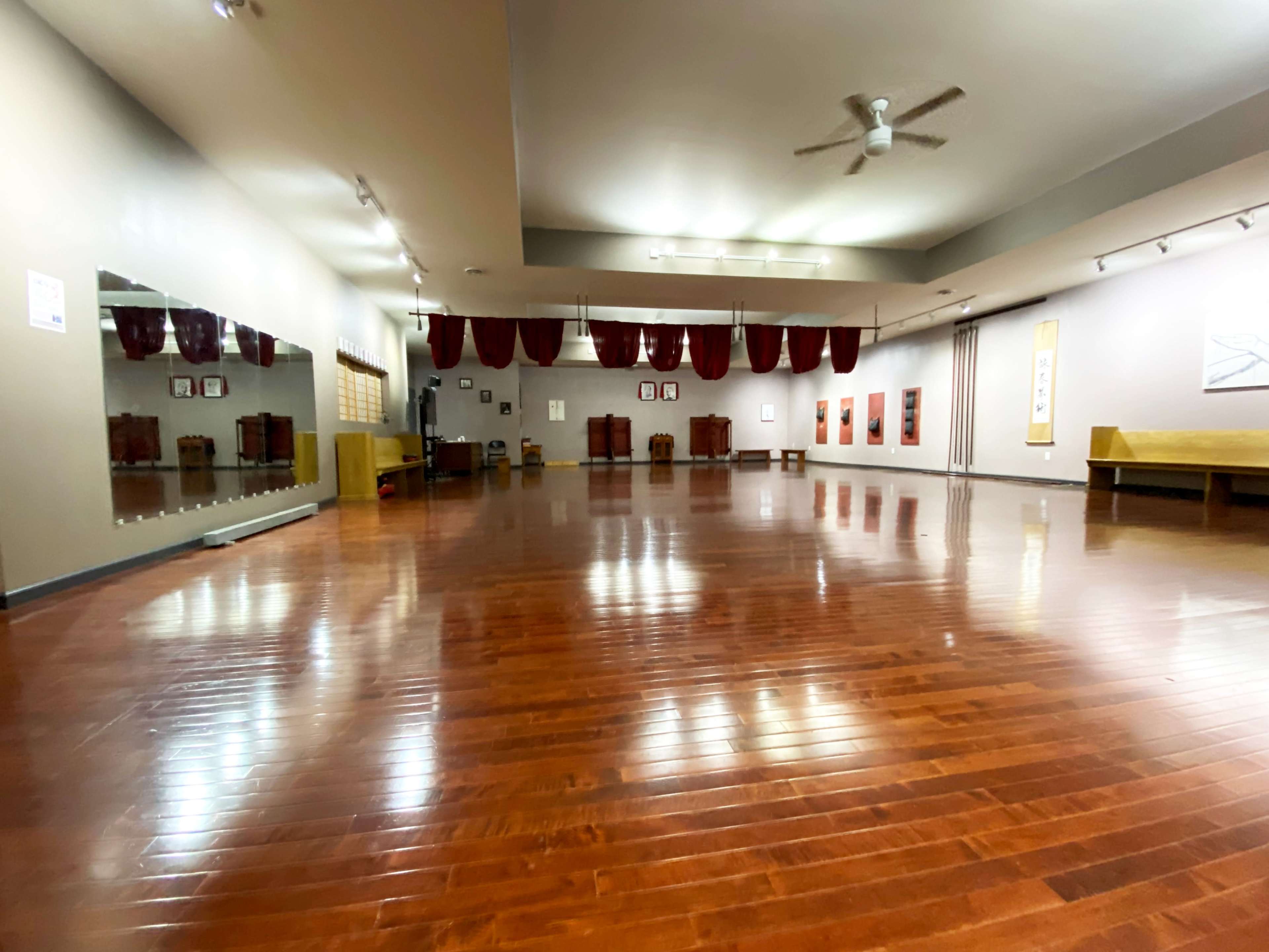 The image shows a spacious, empty studio with polished wooden floors, a large mirror, and red drapes along the ceiling.