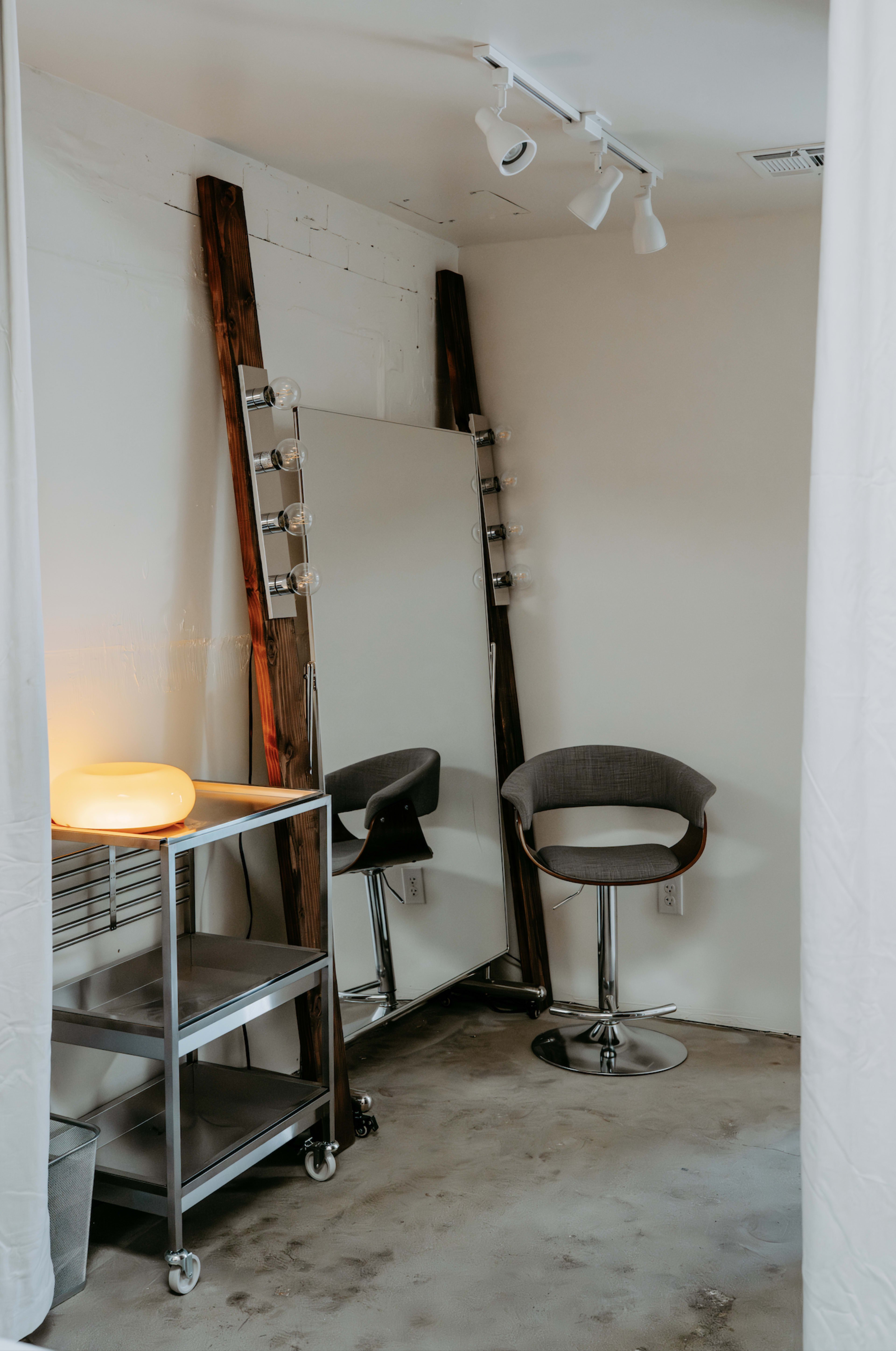 The image shows a modern dressing area featuring a large mirror with lights, two bar stools, a metal cart, and a warm lamp in a minimalistic room.