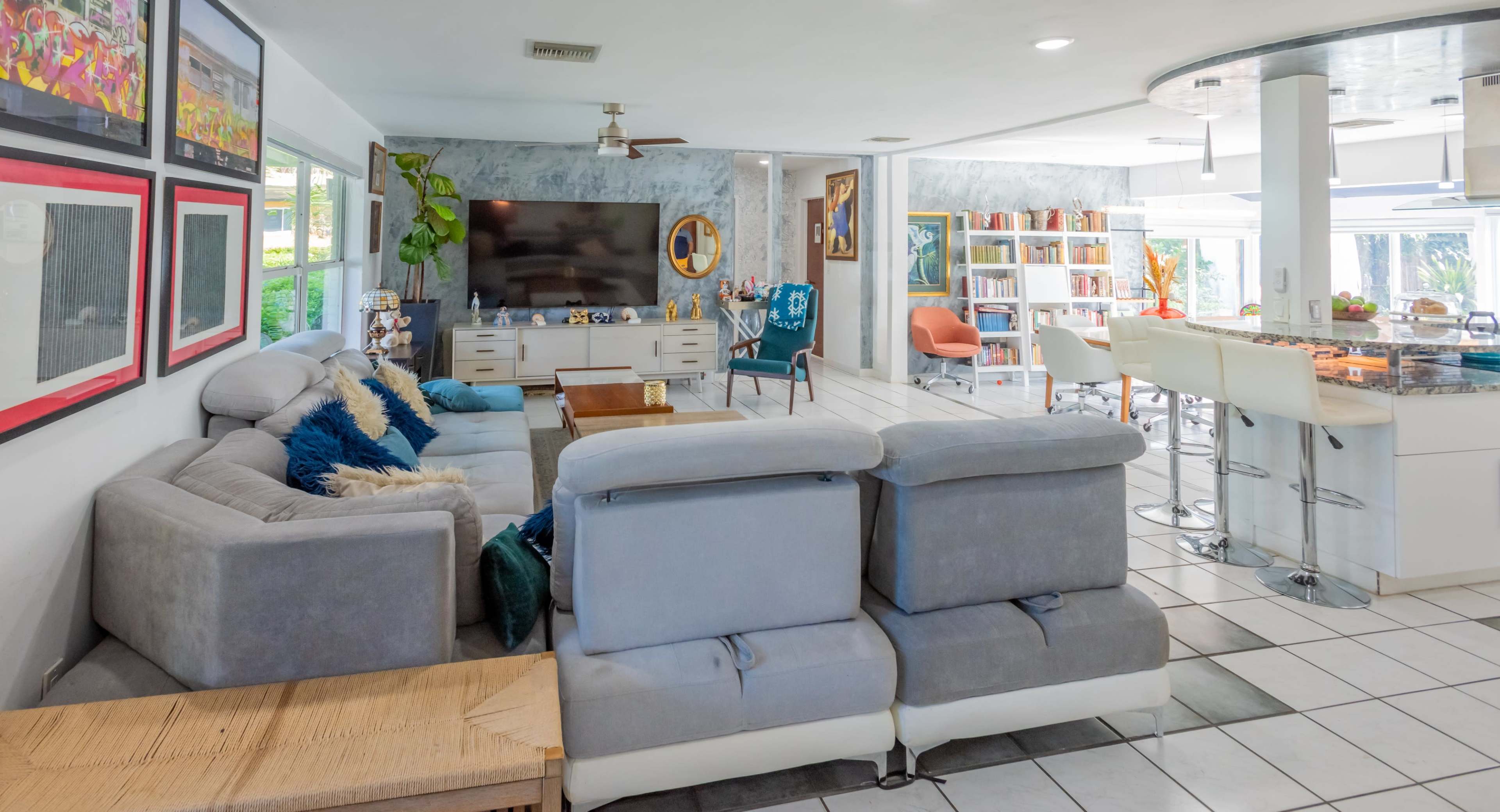 The image shows a modern living room with a gray sectional sofa, a large TV on the wall, and a kitchen area with bar stools and shelves filled with books.