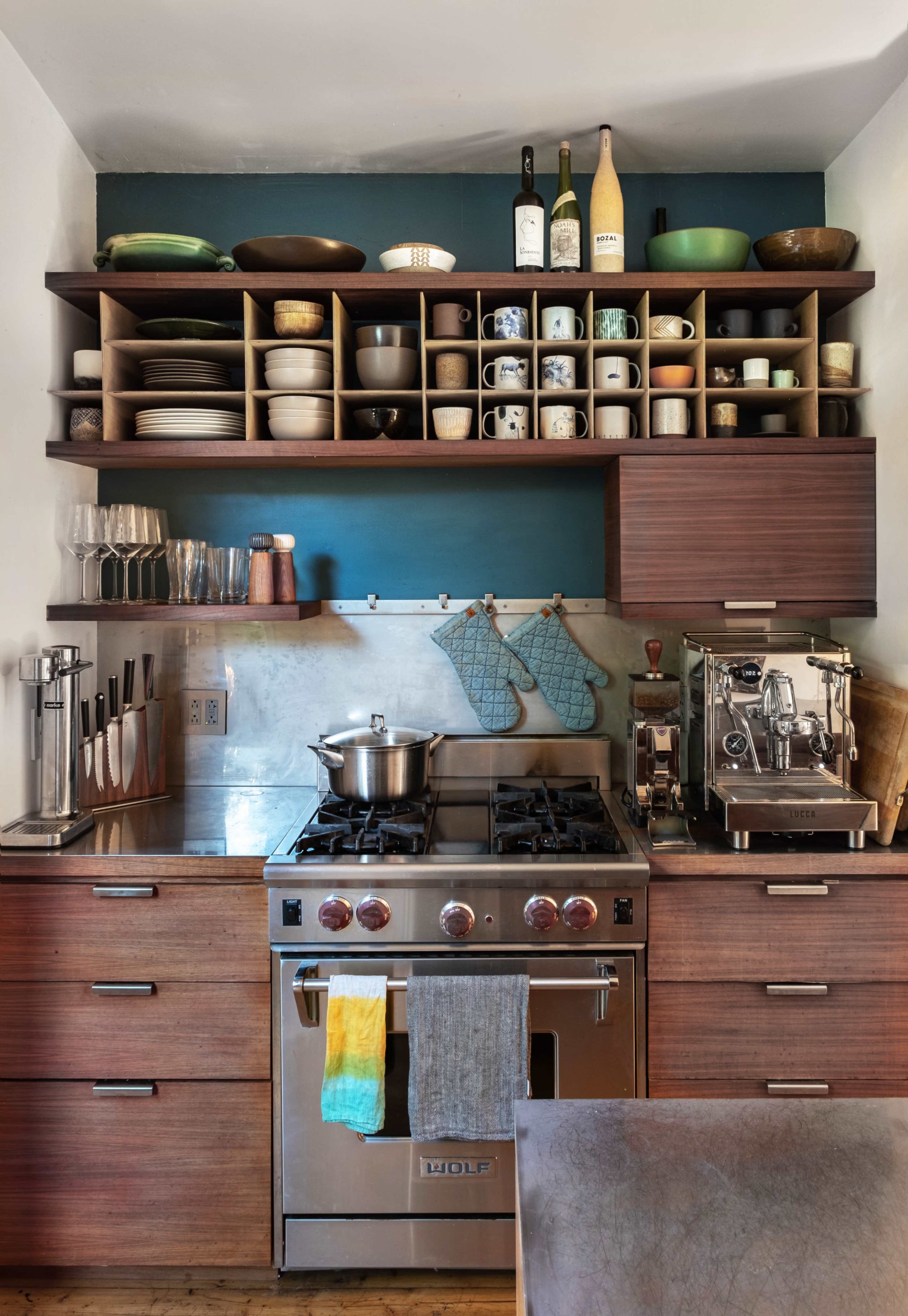 The kitchen features a stainless steel stove beneath a shelf organized with various dishes, glassware, and cooking utensils, complemented by a coffee machine and a pot on the counter.