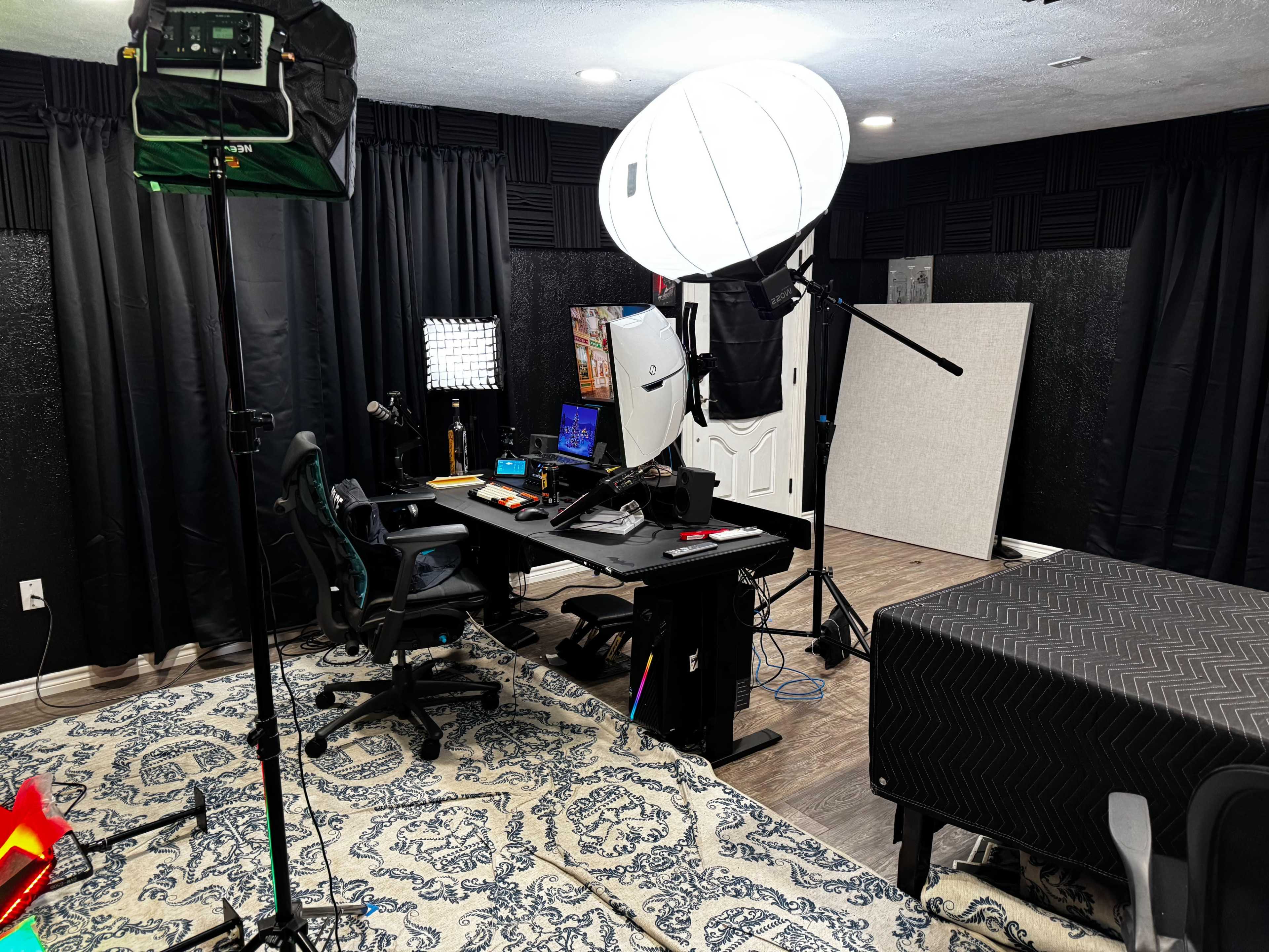 The image shows a well-equipped recording studio featuring a desk with monitors, chairs, and various lighting equipment set against a backdrop of dark curtains and a patterned rug.