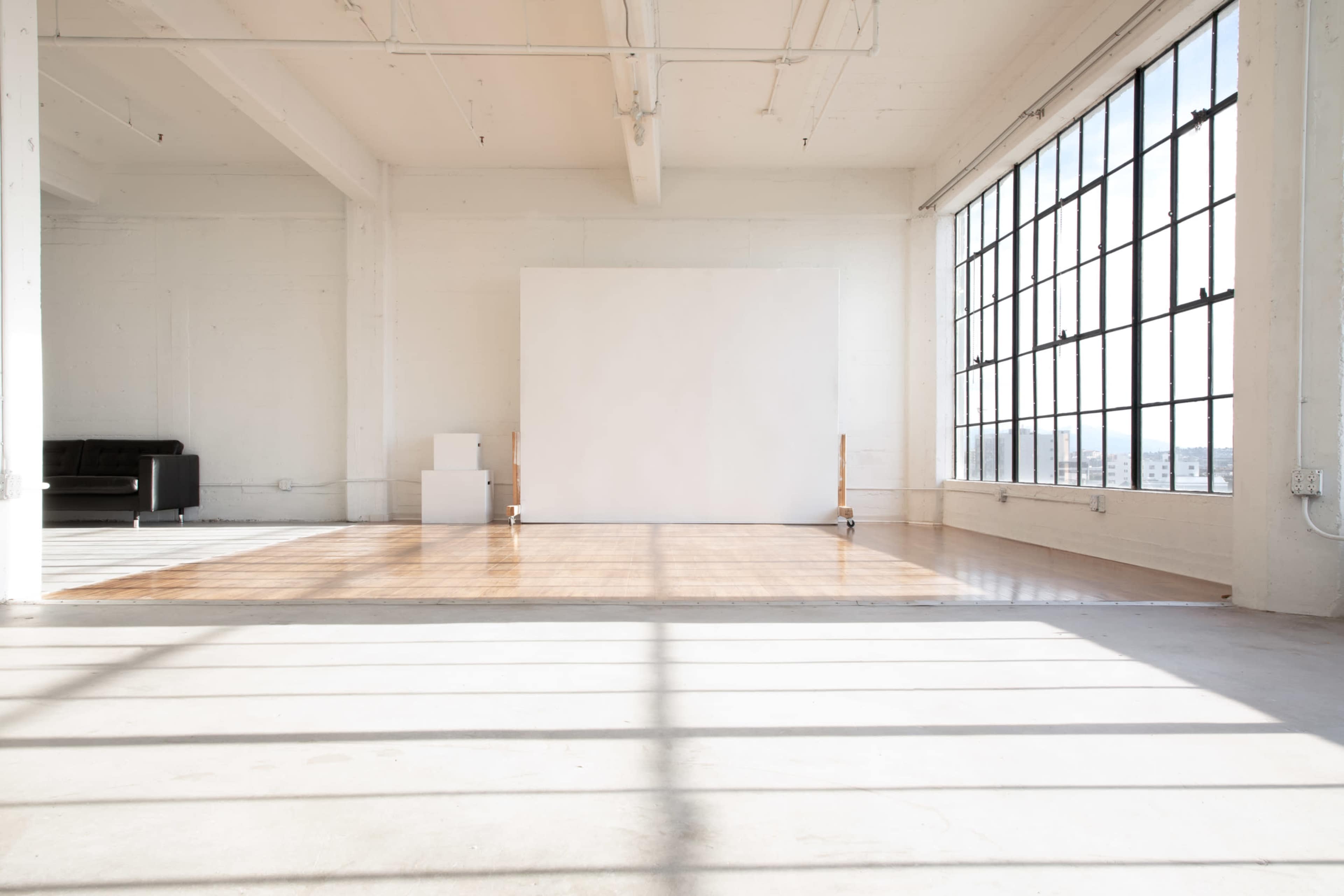 A spacious, bright room with large windows, featuring a polished wooden floor and a blank white wall at one end.