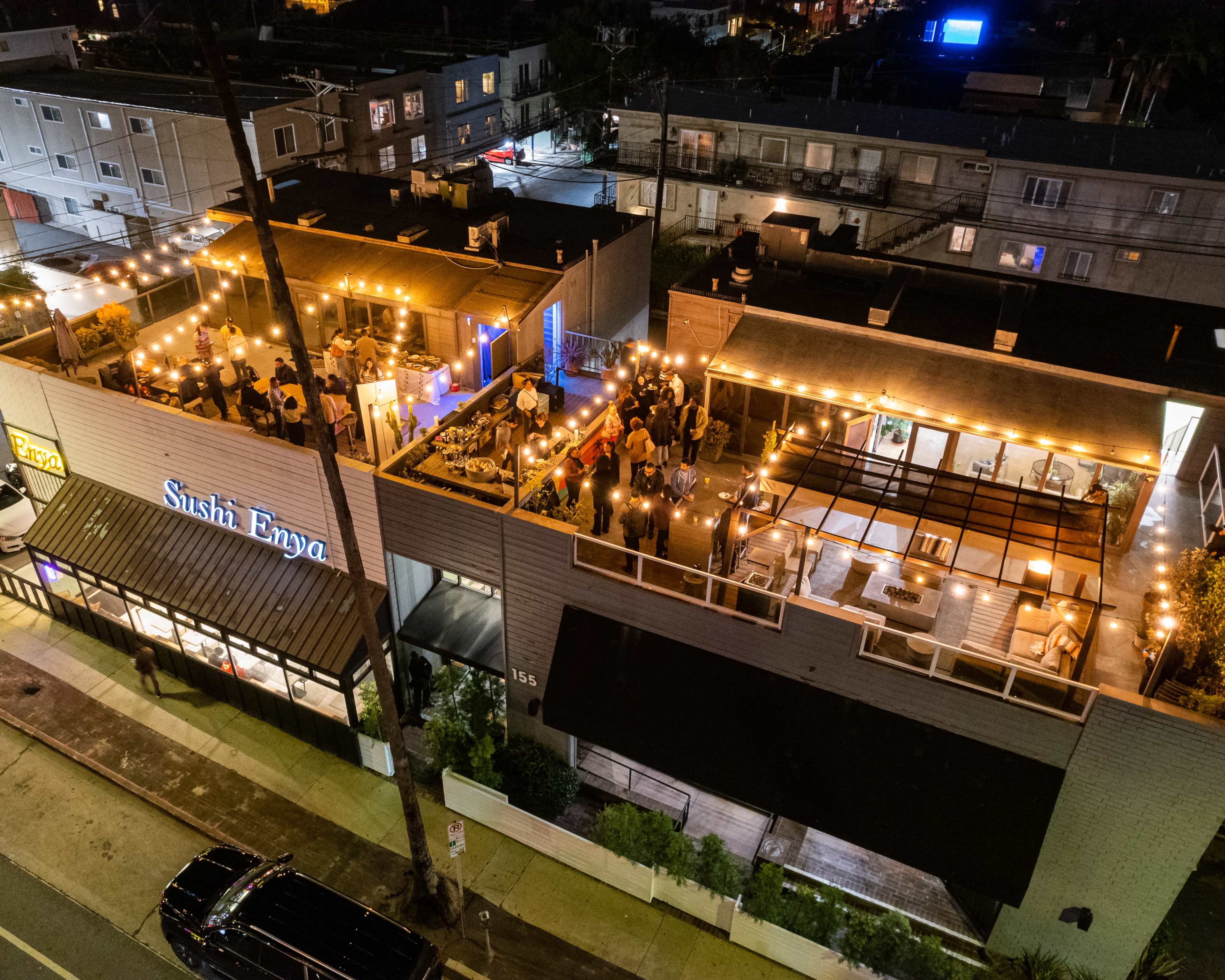 The image shows an outdoor rooftop event at a restaurant called Sushi Enya, with numerous guests mingling under string lights.