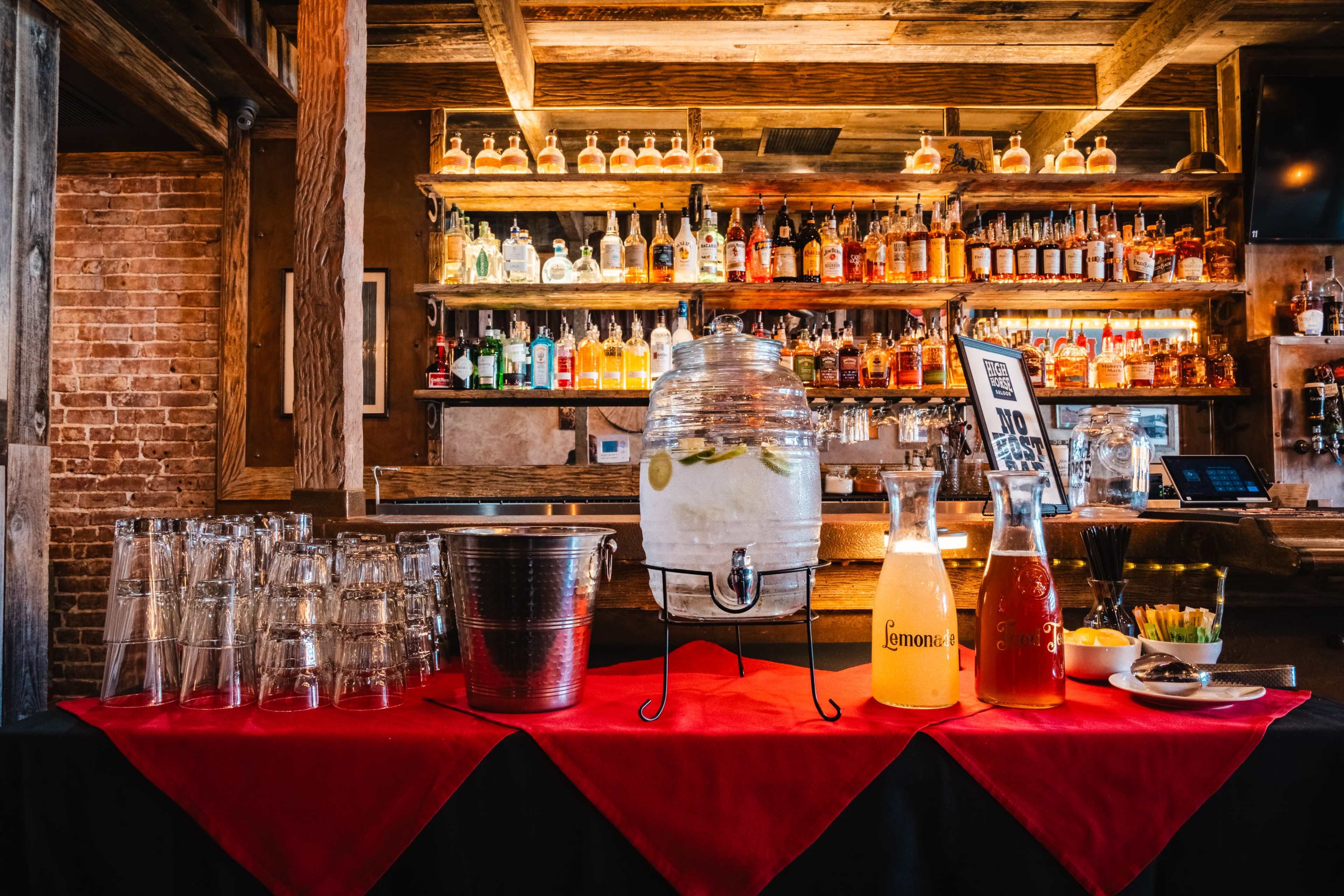 A wooden bar displays an array of colorful liquor bottles on shelves, with a glass container of lemonade and additional beverages on a red-covered table in the foreground.