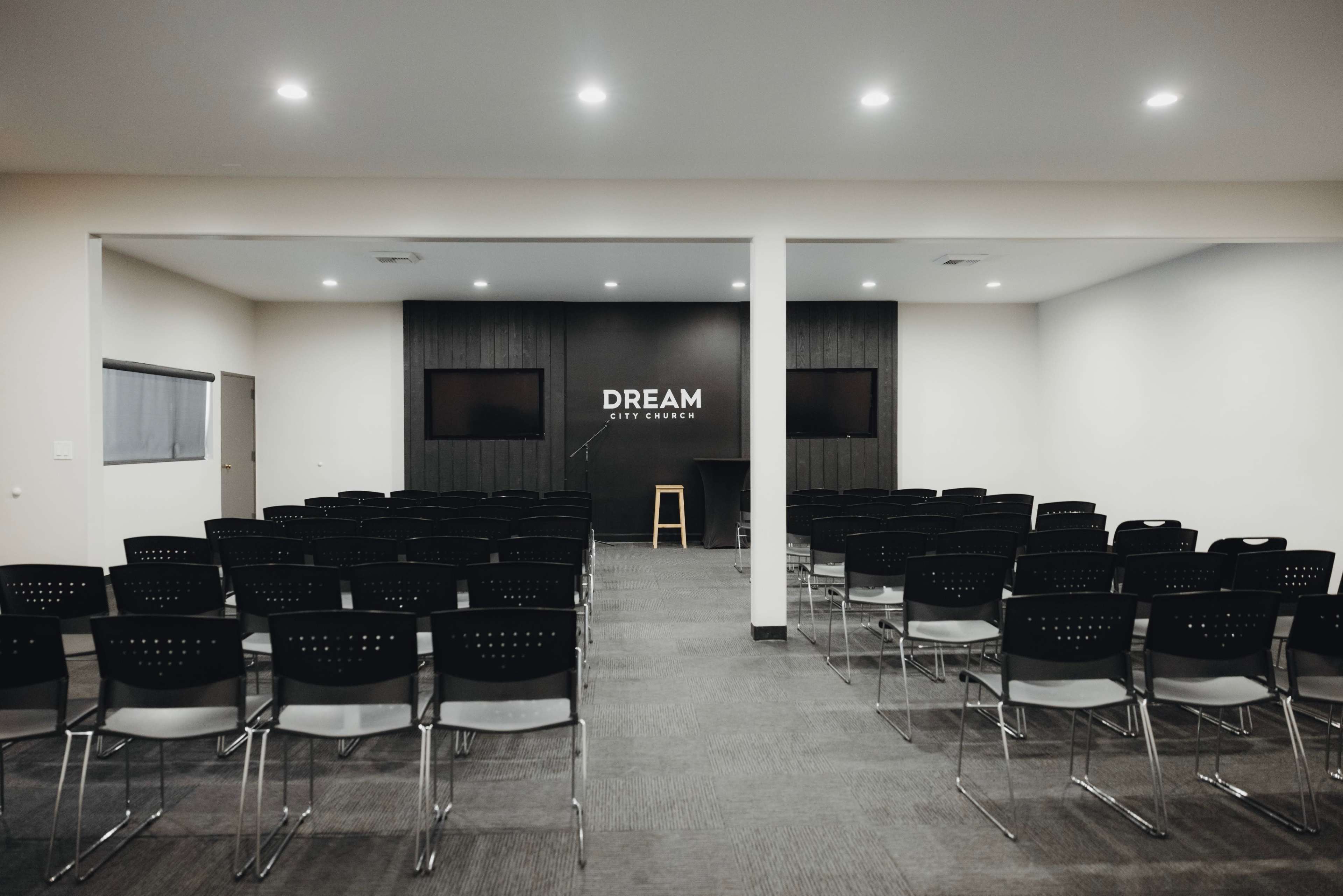A spacious meeting room with rows of black chairs, a wooden stage, and a large sign that reads "DREAM CITY CHURCH" on the back wall.