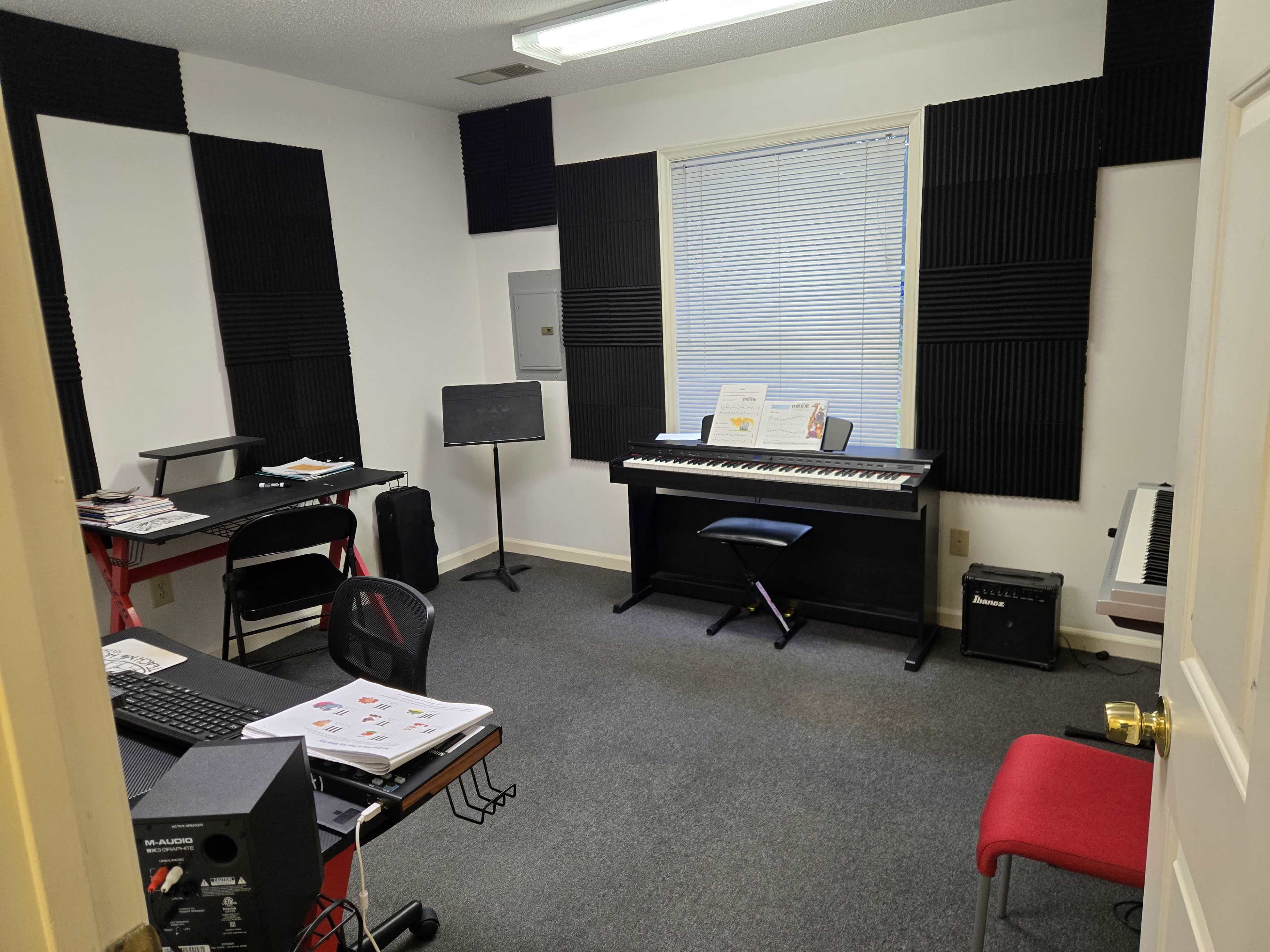The image shows a small music studio equipped with a digital piano, a music stand, and a computer desk, with acoustic panels on the walls.