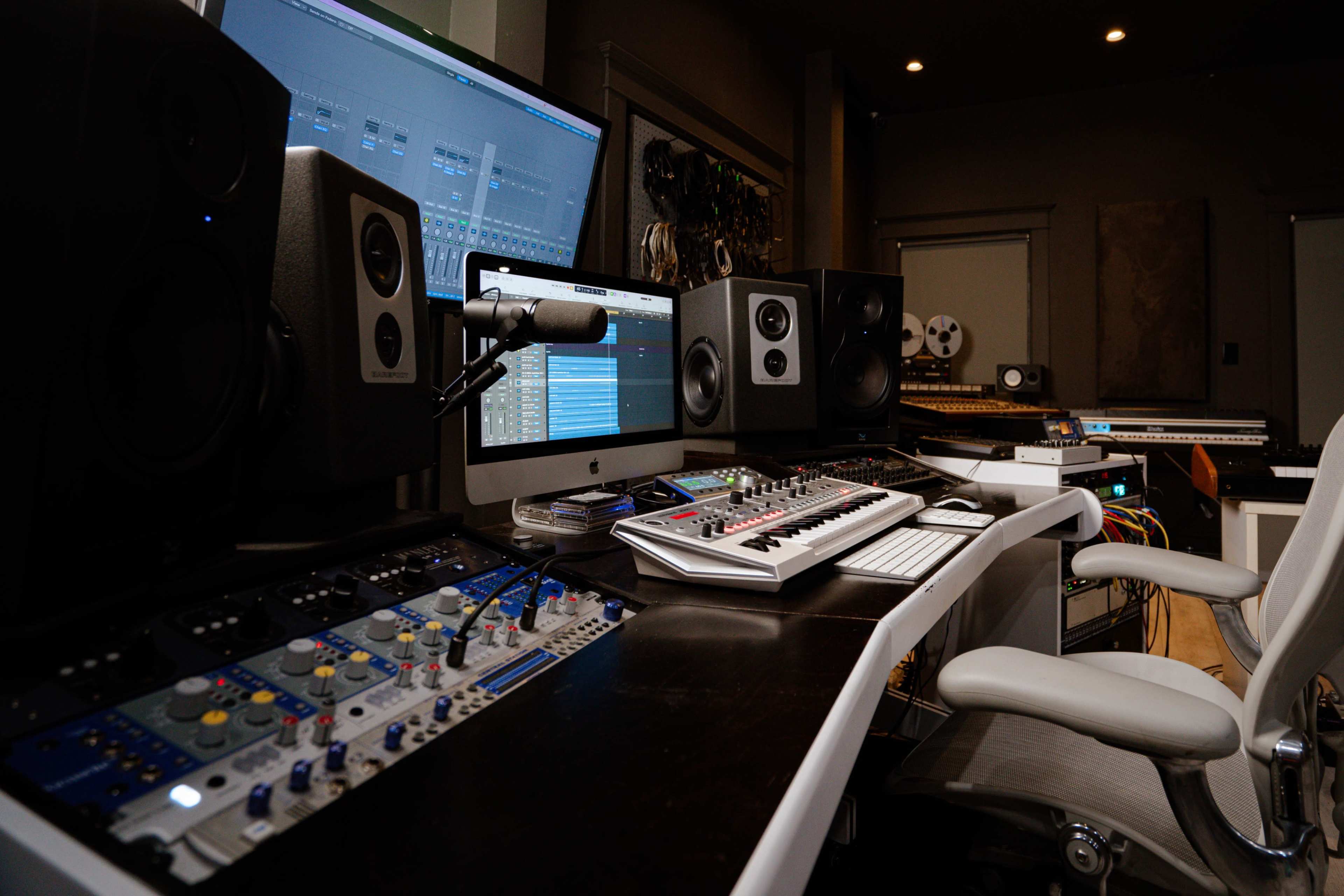 The image shows a modern music production studio with a computer, audio equipment, and various musical instruments neatly arranged on a desk.