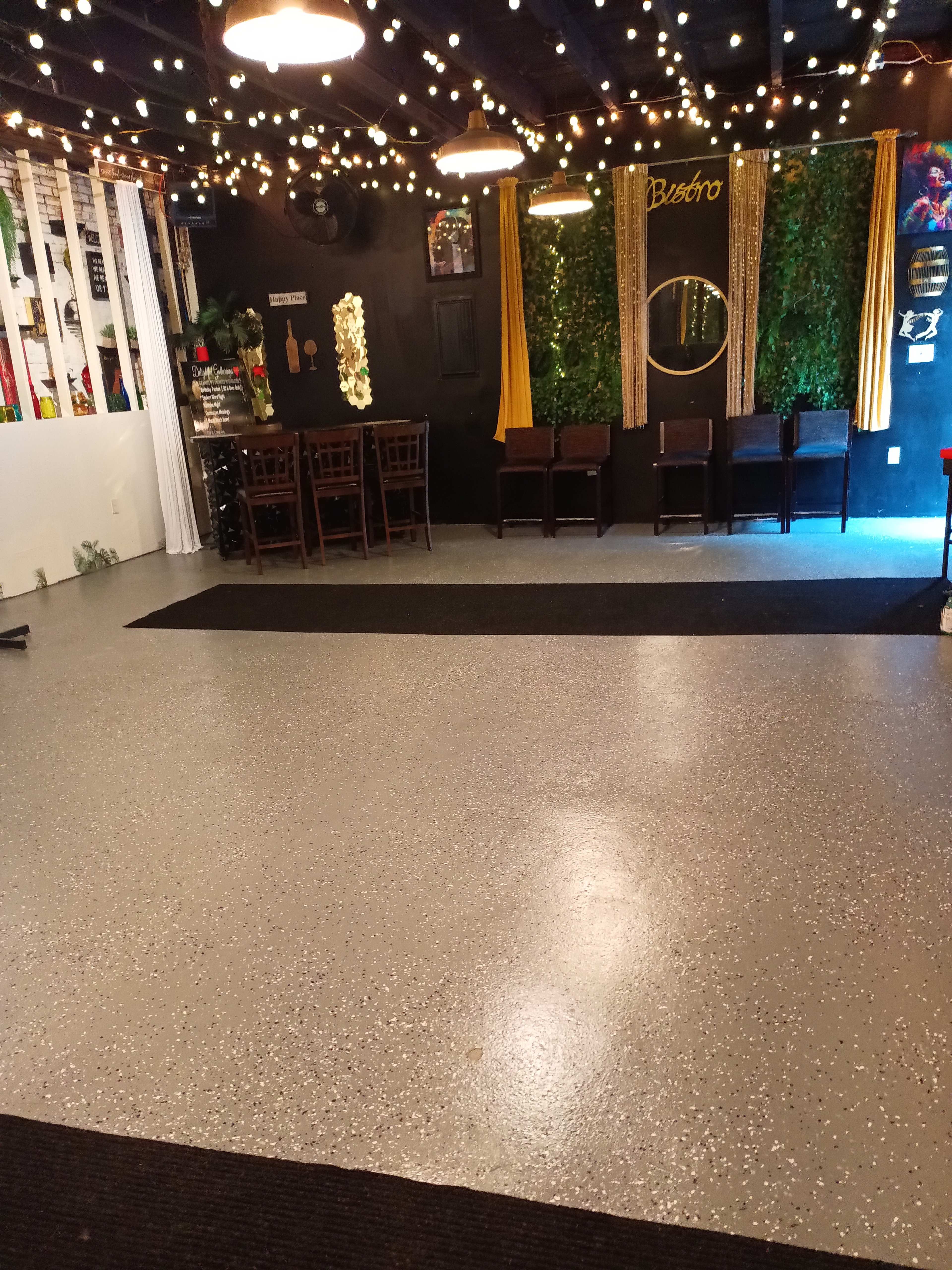 The image shows an interior space with a polished gray floor, string lights overhead, and a seating area featuring wooden chairs and tables against a black wall adorned with greenery.