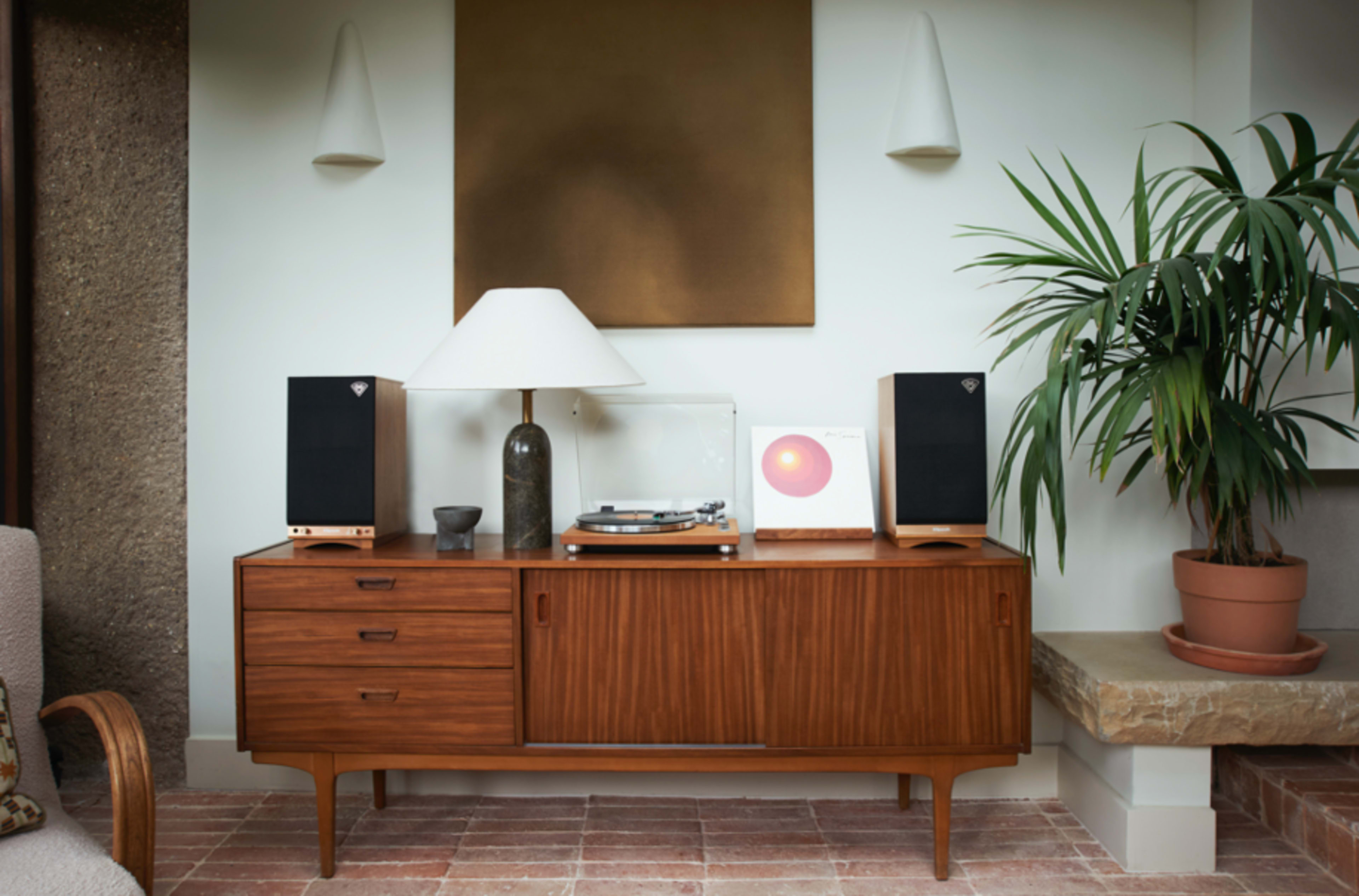 The image shows a mid-century modern wooden sideboard with a turntable, speakers, a lamp, and a vinyl record displayed next to a potted plant.