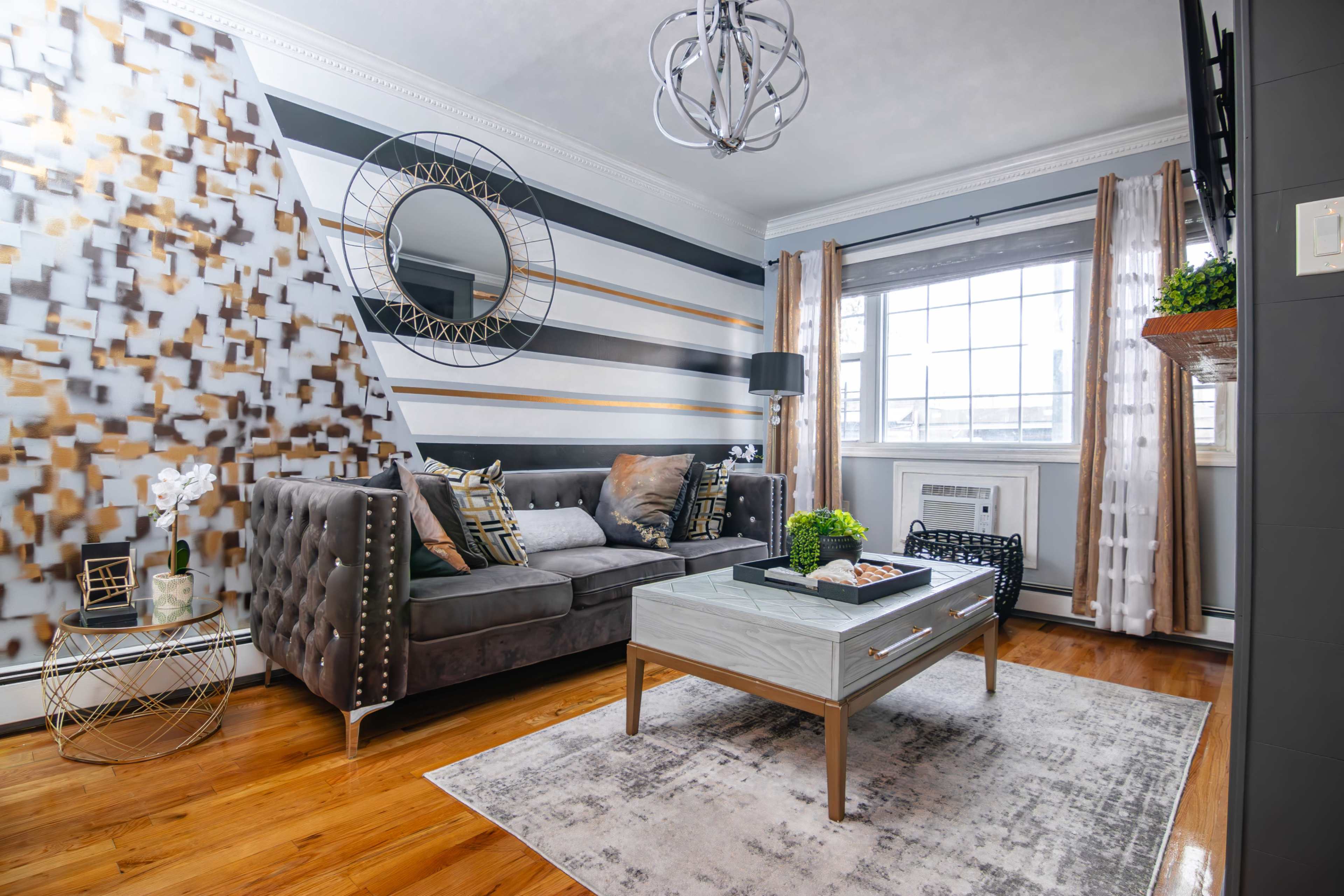 A modern living room with a tufted gray sofa, a mirrored coffee table, and a decorative wall combining geometric patterns in black, white, and gold.