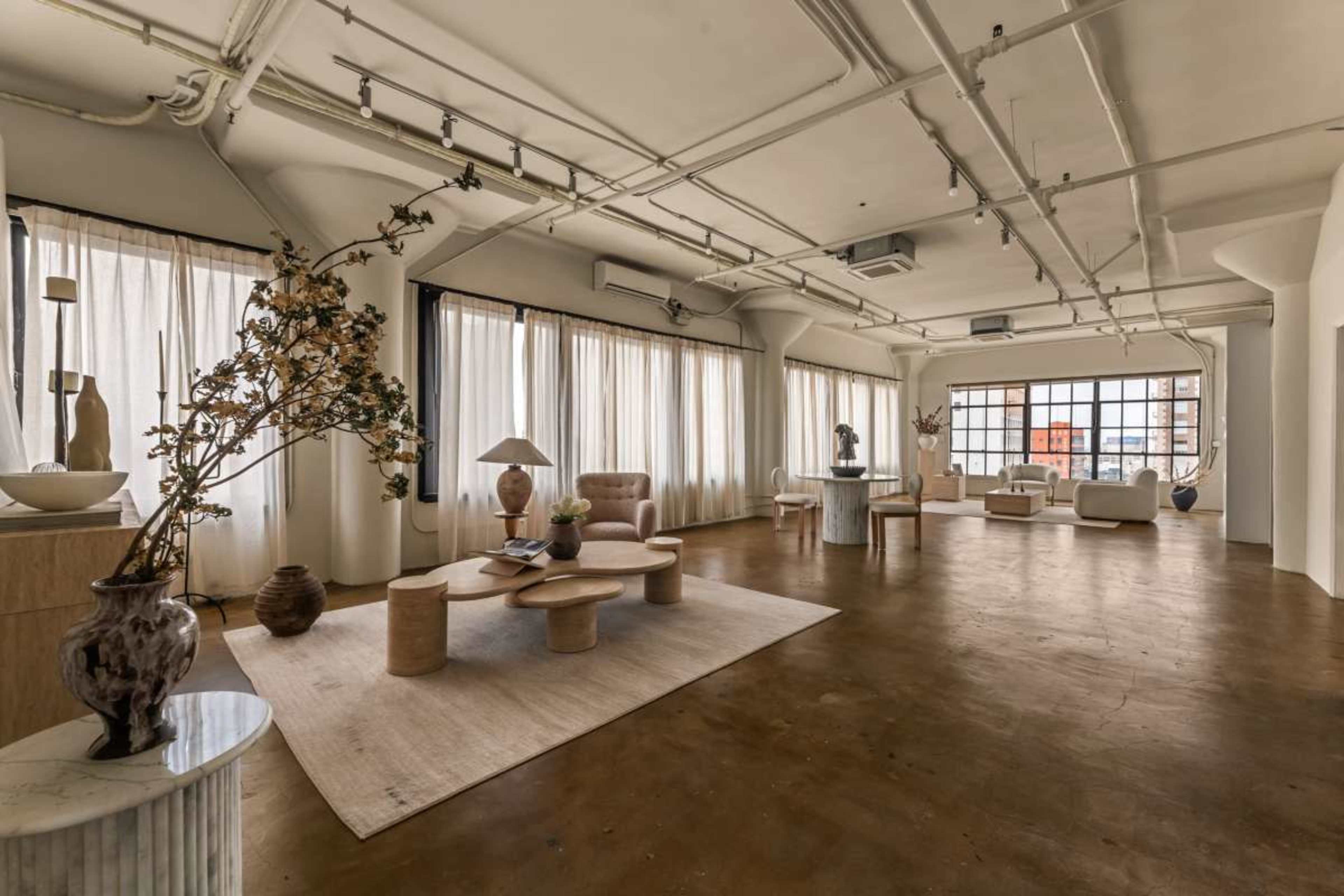 The image shows a spacious, minimalistic interior with large windows, natural light, and contemporary furniture arranged around a central rug.