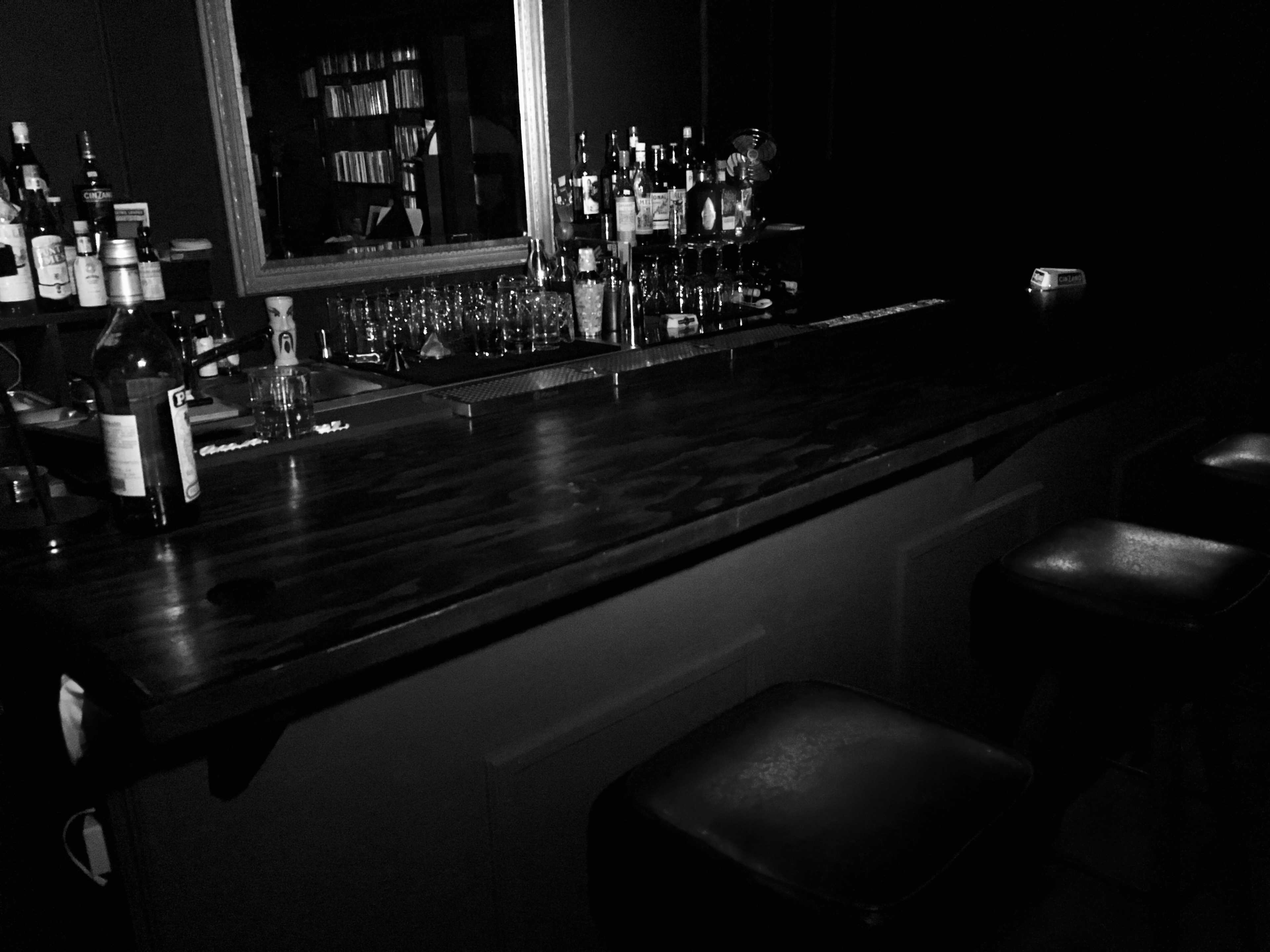 A dimly lit bar scene features a wooden counter lined with bottles and a mirror reflecting the shelves behind.
