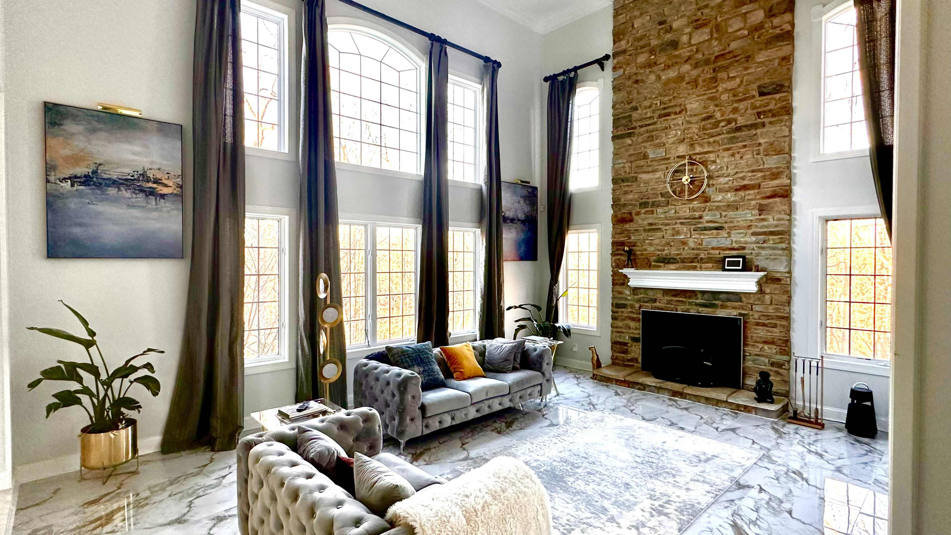 A spacious living room features tall windows, a stone fireplace, and elegant furniture arranged on a marble floor.