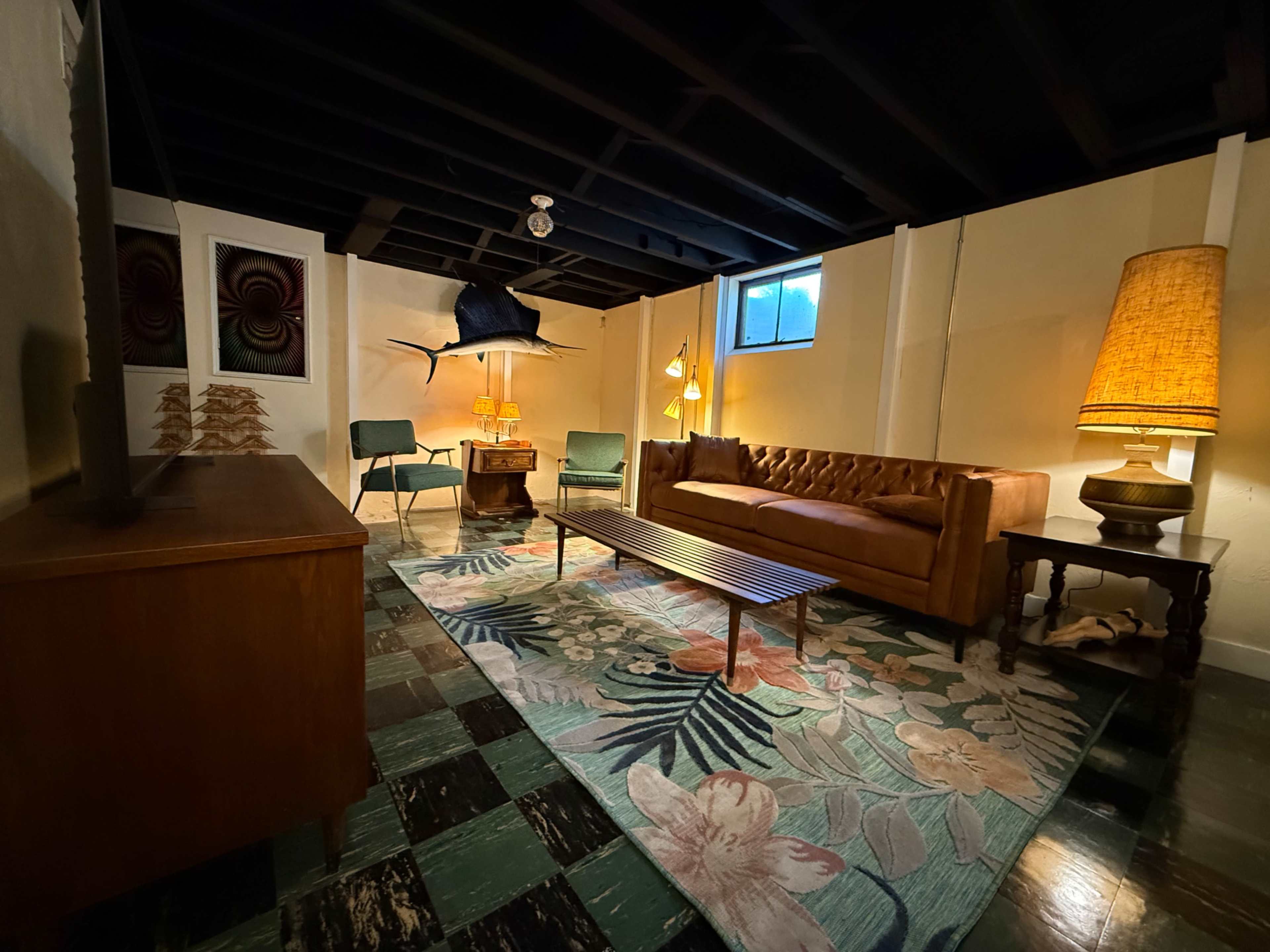 The image shows a dimly lit basement lounge featuring a brown tufted sofa, a large floral area rug, two green chairs, a coffee table, and decorative lamps.