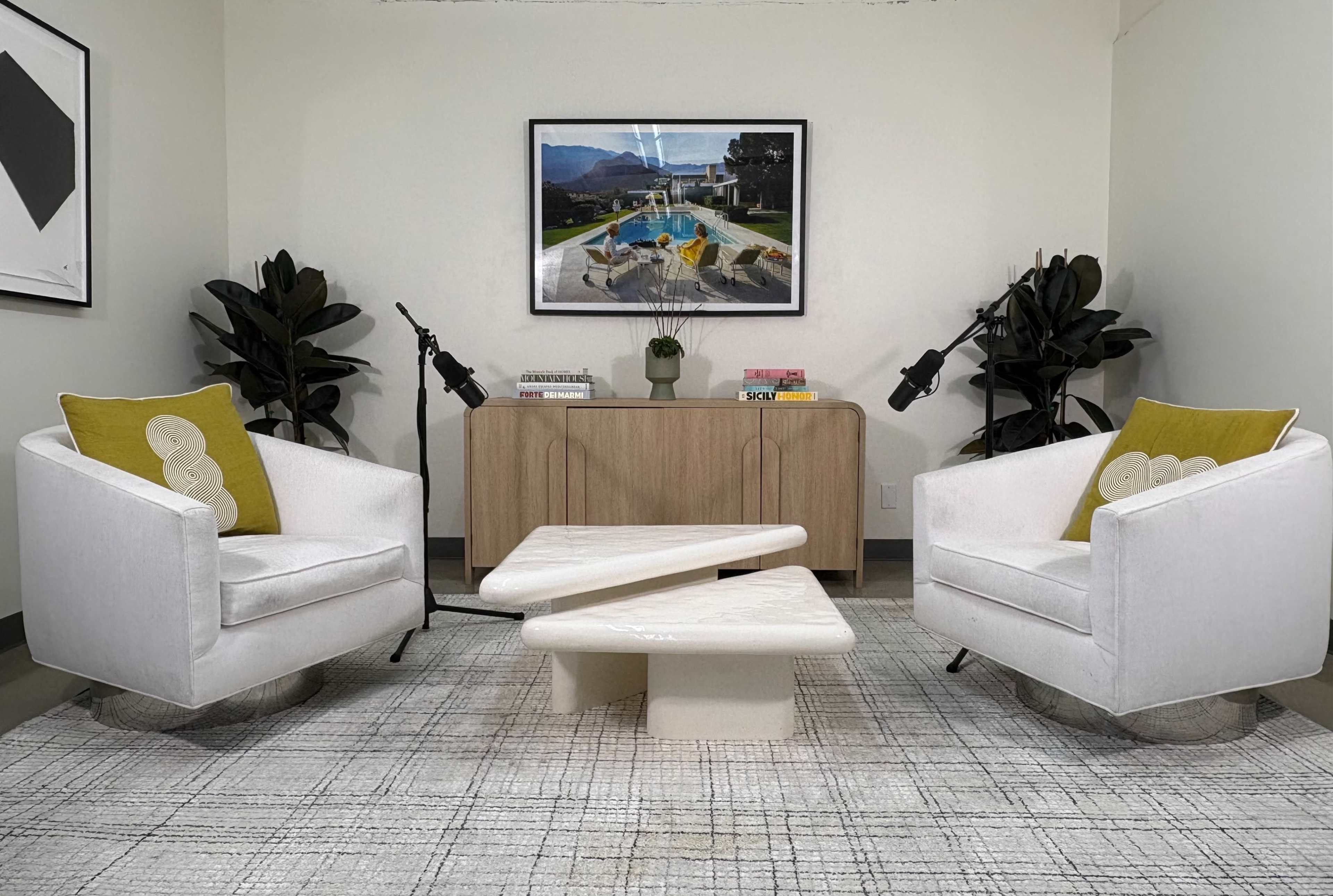 The image shows a contemporary living space with two white armchairs, a sculptural coffee table, a wooden sideboard, and decorative plants, all arranged on a patterned rug.