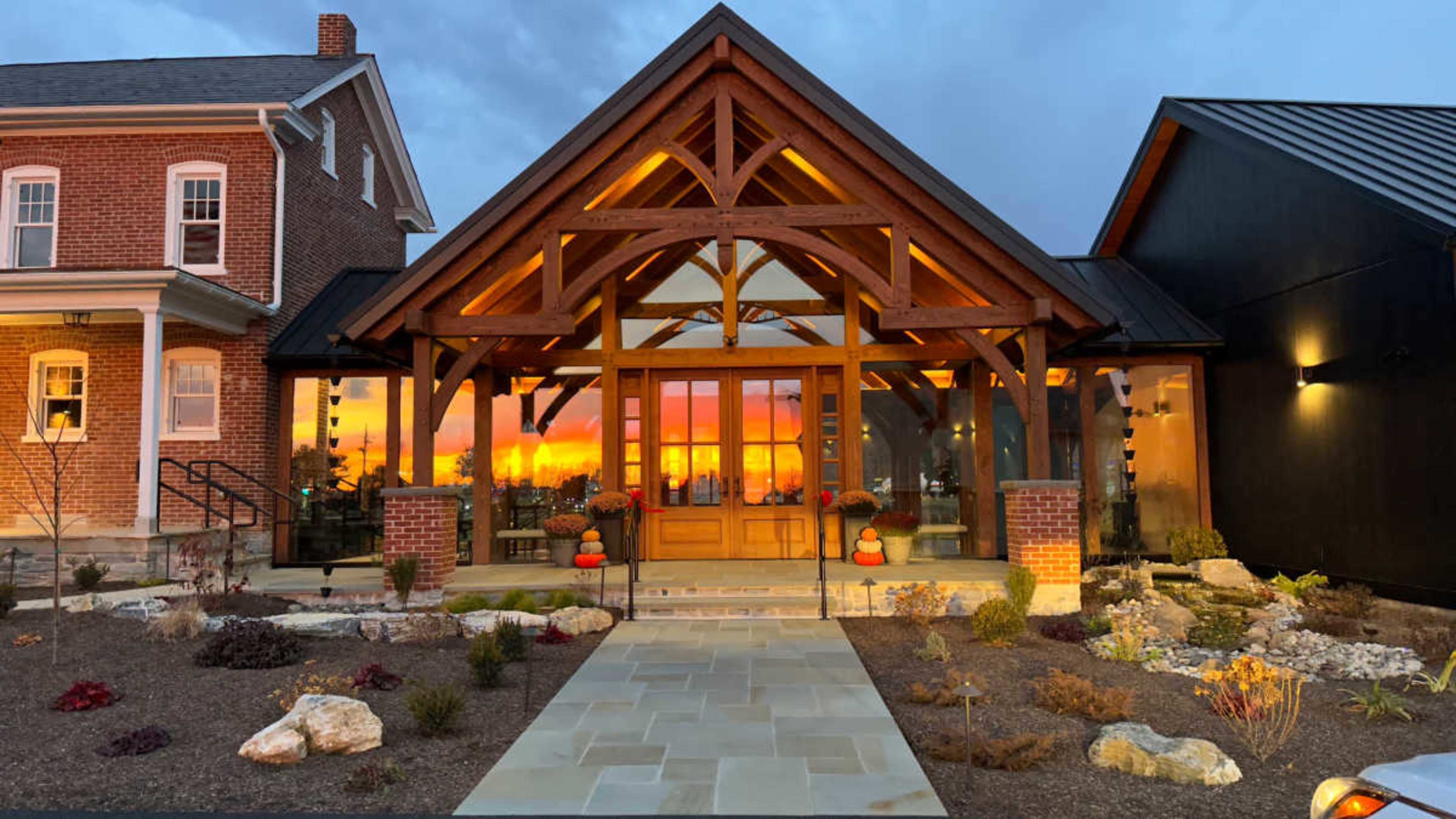 A modern wooden entrance with large glass windows, flanked by gardens and a colorful sunset in the background.
